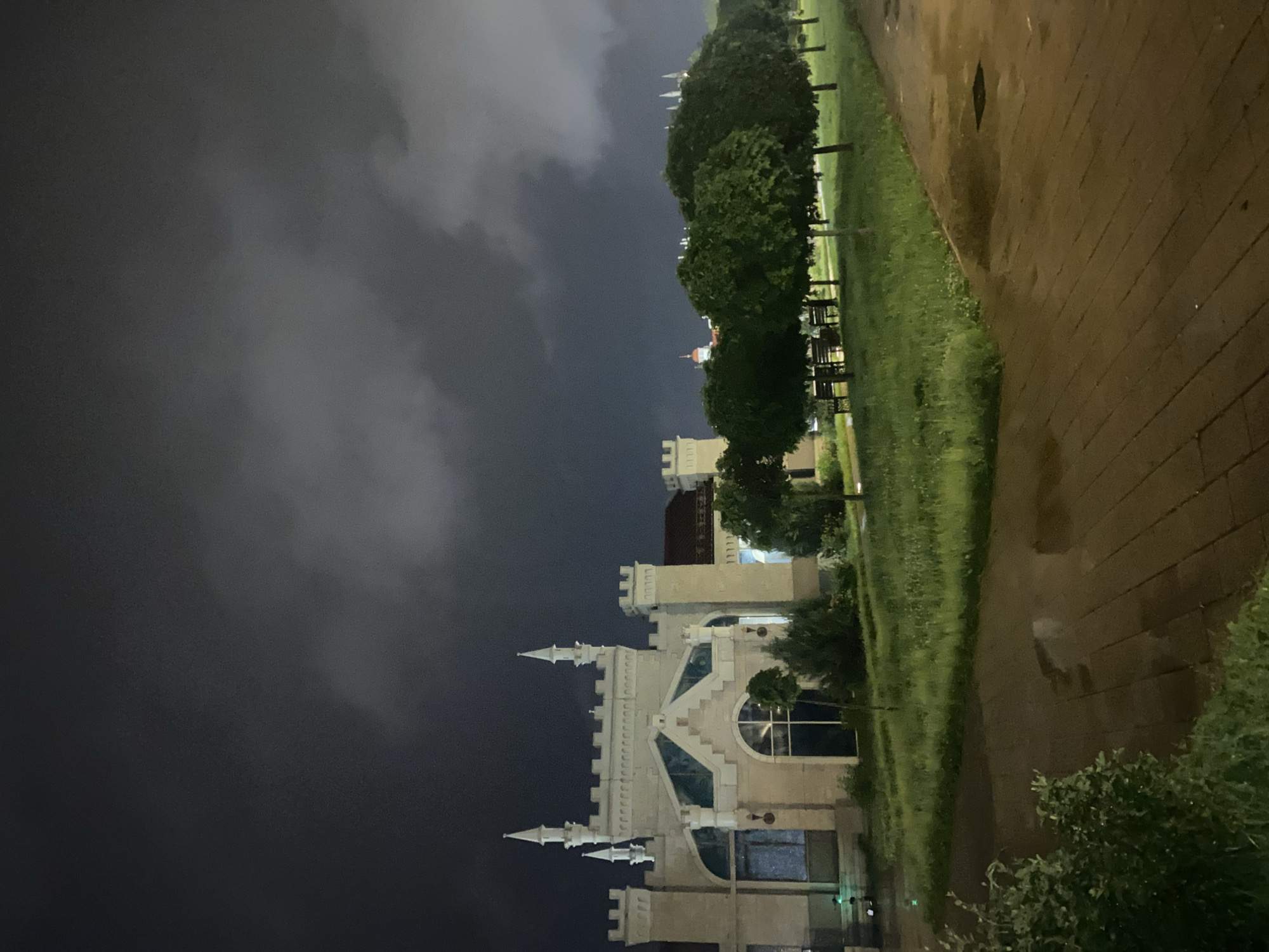 The image captures an exterior view of a building with Gothic-inspired architecture, resembling a castle or a historical manor. The building is illuminated by artificial lighting, highlighting its white facade, turrets, and arched windows. In front of the building, there is a well-maintained lawn with scattered trees, and to the right, a paved walkway with puddles of water, suggesting recent rain. The sky is dark and cloudy, indicating nighttime or overcast weather. In the distance, other buildings and structures are visible. The overall scene conveys a sense of stillness and an almost ethereal ambiance due to the lighting and weather conditions. This specific building is likely the "Ostrov Palace" or "Ostrov Estate" located in Moscow, Russia, known for its distinctive architectural style. There are no people visible in the image.