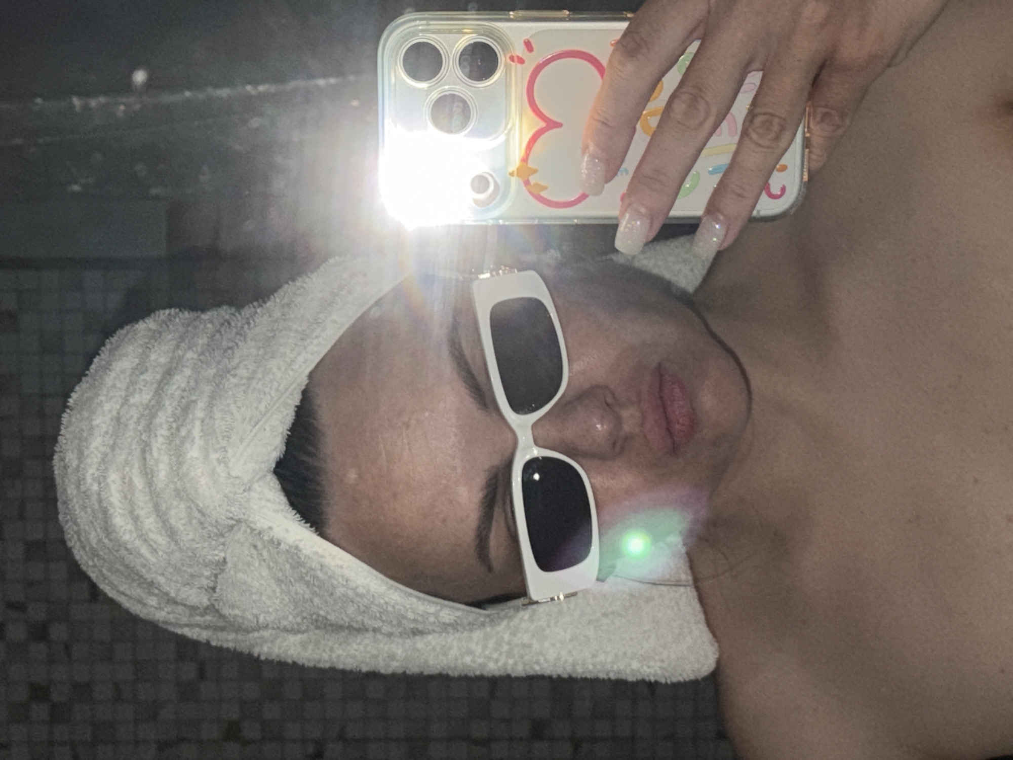 A woman wearing a white towel wrapped around her head and white framed sunglasses is taking a selfie with an iPhone. The flash from the phone reflects off the sunglasses. The woman's chest and shoulder are bare. The background is tiled. There is no indication of the location being in Votkinsk, Russia.