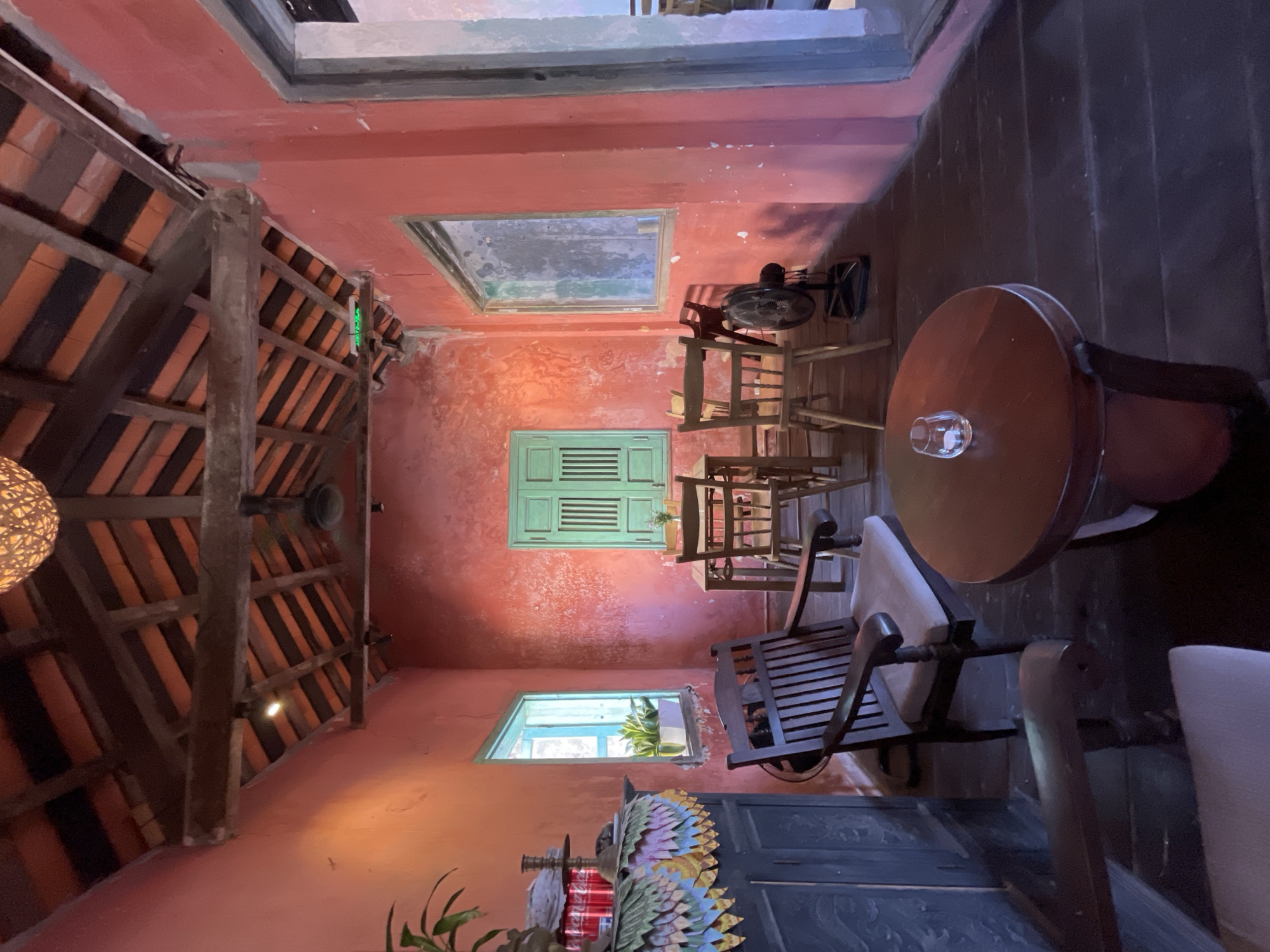 The image depicts the interior of a building in Hội An Ward, Vietnam. The building's walls are painted in a reddish-orange hue. A wooden-beamed ceiling with terracotta tiles dominates the left side of the image.
Several wooden chairs and a round wooden table occupy the right side of the room. A glass sits atop the table. Three windows are visible: two rectangular ones with greenish frames and one larger rectangular window near the ceiling. A standing fan is positioned near the largest window.
In the lower part of the image, there is a dark-colored cabinet with a decorative element on top, featuring what appears to be a depiction of plumage. A plant is visible in the lower left corner.