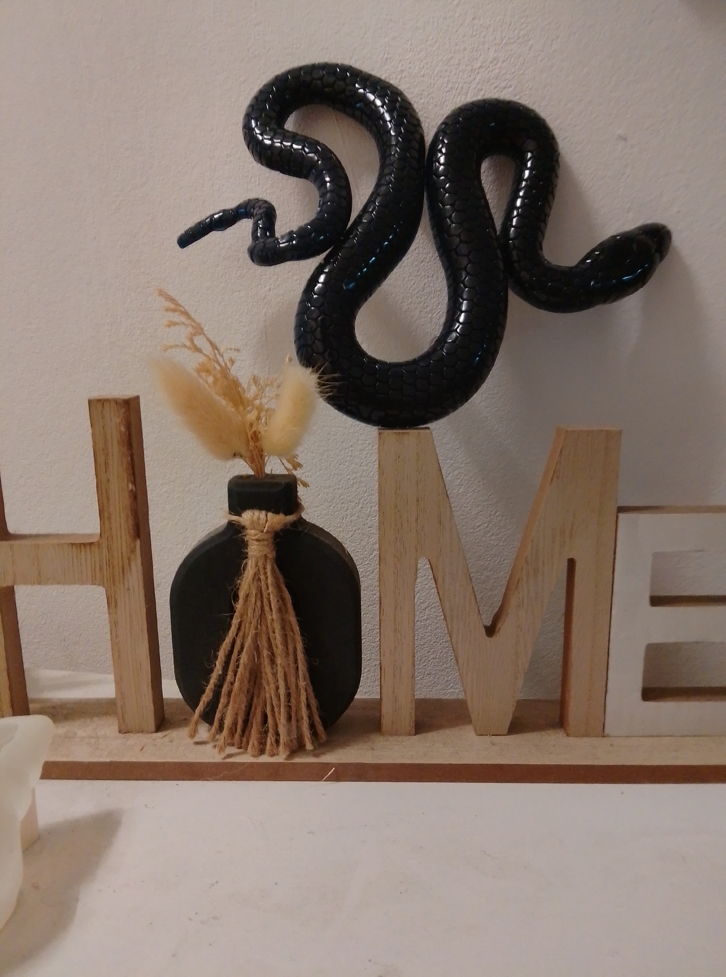A black, glossy, coiled snake sculpture is mounted on a textured white wall. Below the snake, a wooden base displays three large wooden letters, forming the word "HOME". The letter "H" is on the left, followed by a black vase with dried beige ornamental grass and jute twine tied around its neck. The letters "O", "M", and "E" complete the word. The "M" and "E" are made of light-colored wood, while the "E" has a white insert. The overall scene is a decorative display, possibly for a home interior. No people are visible, and the setting appears to be indoors, likely a room in a house. There are no clear indicators of time of day or weather. The location context provided is Wedel, Germany, but there are no visual cues within the image to confirm this.