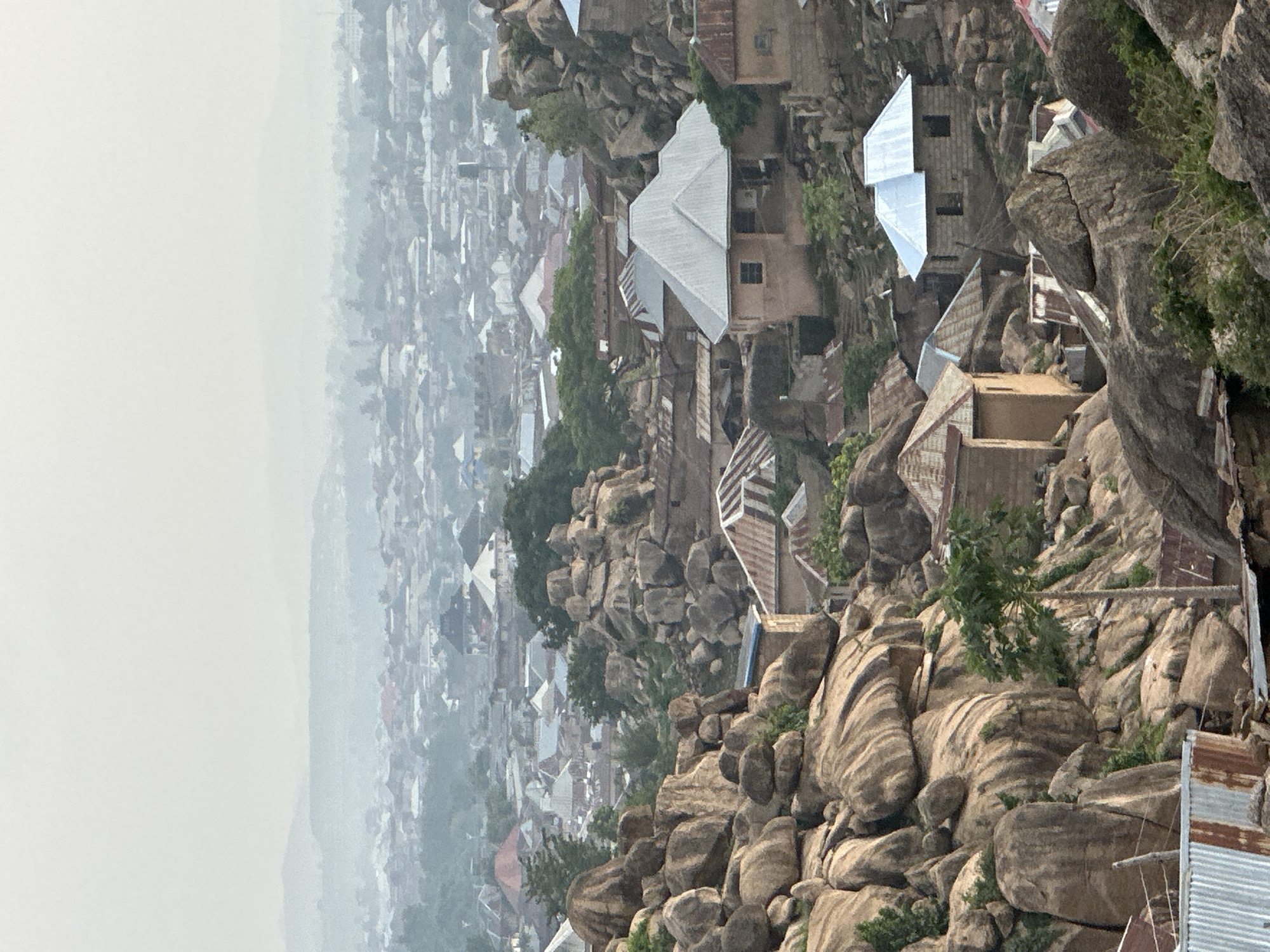 This image captures a panoramic, high-angle view of a densely populated urban or semi-urban area in Jos, Nigeria, distinctly characterized by its unique topography. The scene is dominated by numerous large, rounded granite rock formations (inselbergs or kopjes) that are seamlessly integrated into the settlement.
In the foreground and midground, a multitude of buildings, primarily residential structures, are constructed directly on, around, and between these prominent rocky outcrops. Many of these buildings feature distinctive corrugated metal roofs, varying in color from silver-grey to rust-brown, indicating age or material differences. The architecture appears functional and adapted to the rugged terrain, with some structures seemingly built into the rock faces themselves. Sparse green vegetation, including trees and bushes, dots the landscape, growing amidst the rocks and around the dwellings.
As the eye moves towards the background, the settlement stretches into the distance, forming a vast expanse of tightly packed structures. The visibility progressively decreases due to a pervasive haze, which blankets the distant cityscape and obscures the horizon, blending the sky with the furthest buildings. This atmospheric condition suggests either early morning mist, a humid day, or atmospheric pollution.
The overall impression is one of a vibrant human settlement coexisting with and adapting to a challenging natural environment. No specific human activities or individuals are distinctly visible, giving the scene a quiet, almost paused quality. There is no visible text in the image. The high vantage point offers a comprehensive overview of this unique landscape in Jos, Nigeria, under hazy daytime conditions.