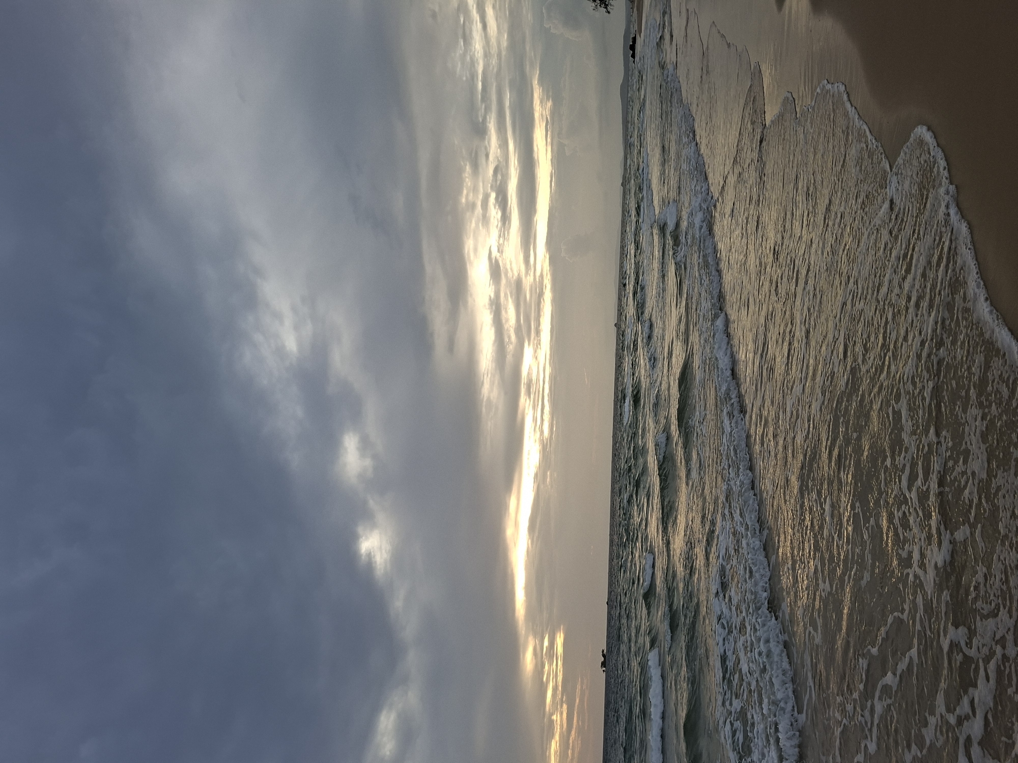 The image depicts a wide shot of a beach with a cloudy sky and calm waves. The sky is filled with dark, moody clouds with streaks of light breaking through, suggesting either sunrise or sunset. The ocean is a deep blue-green with gentle waves rolling onto the shore, creating white foam as they break. The sand is a light brown and appears wet near the water's edge. In the distance, there are a few small boats visible on the horizon, and a silhouette of land can be seen. The overall scene is serene and natural, with no people or specific structures prominently featured. The location is identified as a beach in Phú Quốc, Vietnam.