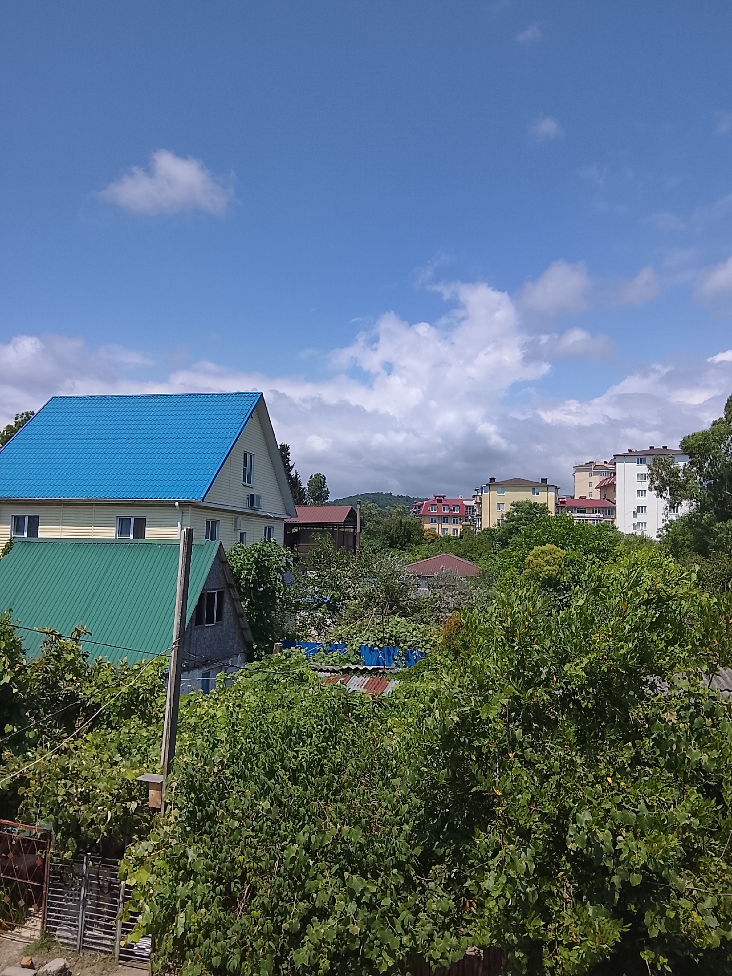The image depicts a residential area on a sunny day with scattered clouds. In the foreground, a dense canopy of lush green trees dominates the scene. Partially visible through the foliage are various structures. A prominent house with a light-colored exterior and a vibrant blue metal roof is situated towards the left. Adjacent to it, a smaller structure with a green metal roof is also visible. Further in the background, several multi-story buildings, likely apartment complexes, rise above the trees, suggesting a developed urban or suburban setting. One of these buildings is white with numerous windows, and others are painted in yellow and red hues. The sky is a clear blue, with a few white, puffy clouds scattered across it. The lighting suggests it is daytime, likely late morning or early afternoon. The overall impression is one of a peaceful, established community bathed in warm sunlight. No text is visible in the image. The location context of Oryol-Izumrud, Russia, indicates this is likely a scene from a town or city in that region. The abundance of greenery suggests a mild climate.