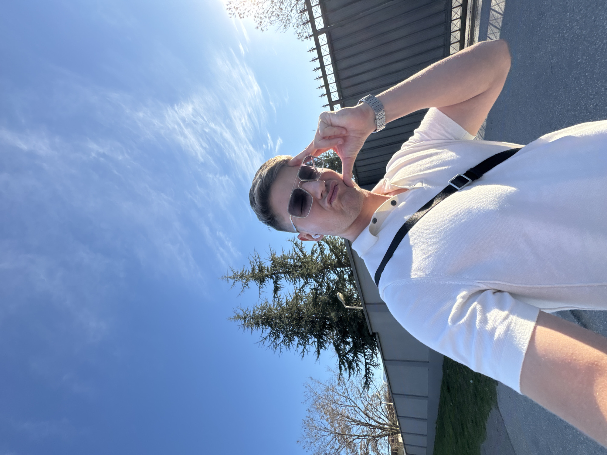 A man poses for a selfie outdoors in Plovdiv, Bulgaria, on a sunny day with a blue sky streaked with cirrus clouds. He is wearing a white polo shirt, sunglasses, and a watch. A black strap is slung across his chest. He is holding his hand up to make a square shape around his eye. Behind him are coniferous trees and a fence.