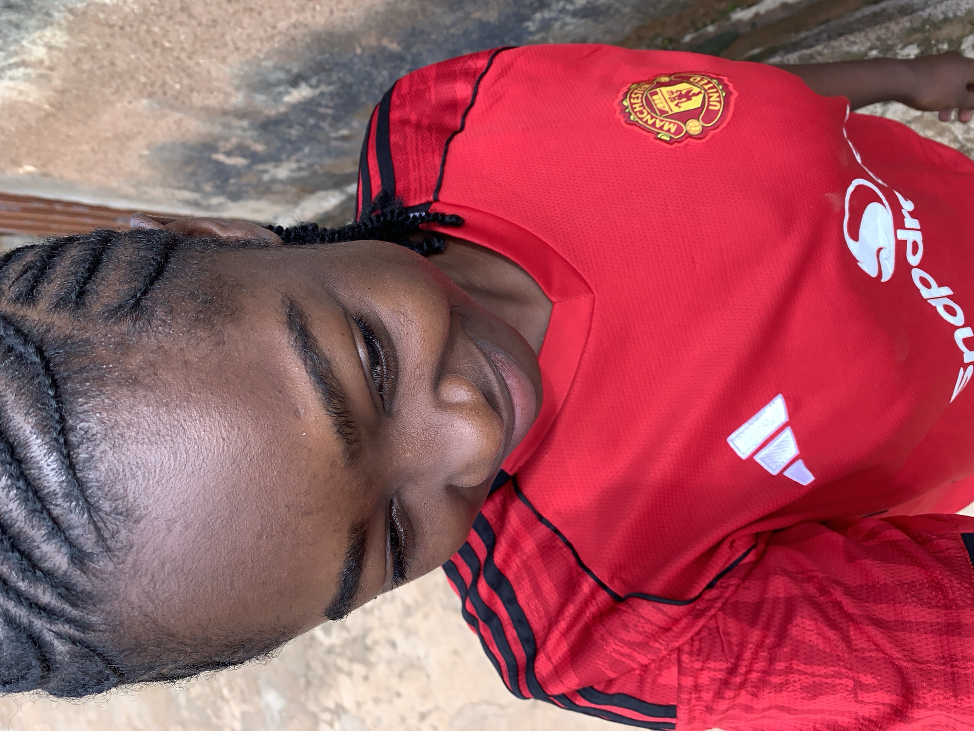 The image captures a close-up, angled portrait of a person, likely a youth or young adult, with dark skin and neatly braided hair (cornrows) visible on the side of their head. The individual's eyes are closed or heavily lidded, and their head is tilted slightly, conveying a sense of serenity or contemplation.
They are wearing a vibrant red football jersey, clearly identified as a Manchester United kit. The jersey features the Manchester United crest on the left chest, displaying a ship and a red devil, with the words "MANCHESTER UNITED" encircling it. On the right side of the chest/shoulder, a white Adidas logo with three distinctive stripes is visible. The primary sponsor's name, "Snapdragon," is printed vertically in white on the lower right side of the jersey. Black Adidas stripes run down the shoulders of the red shirt. The jersey fabric has a discernible texture.
The background is out of focus, showing a mottled, earthy-toned surface that suggests an outdoor setting, possibly a wall or ground, in what is identified as Rafin Par, Nigeria. The lighting indicates it is daytime. The image provides a focused view of the person's features and their sports attire.