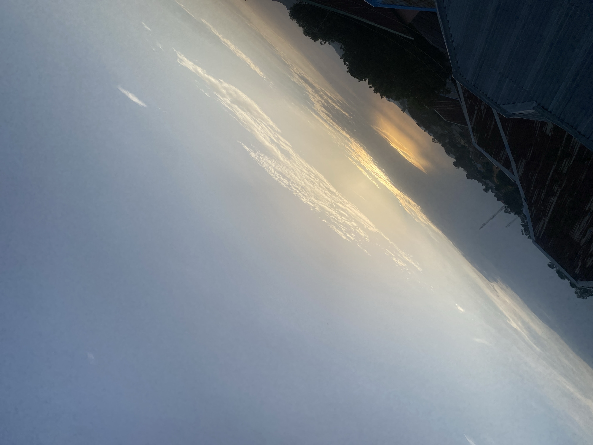 This is a photograph taken outdoors, with the camera tilted. The scene depicts a hazy sky with streaks of clouds, suggesting a possibly humid or smoggy atmosphere. The sun appears to be setting or rising, casting a warm, golden glow through the clouds in the upper right portion of the frame.
In the foreground, a textured, dark surface occupies the right side of the image, possibly a rooftop or a constructed walkway, with distinct lines and panels. Beyond this structure, a dark line of trees forms a silhouette against the hazy sky, indicating land or a hillside. The overall impression is of a natural landscape viewed from an elevated position, possibly during twilight. The specific location is identified as Bukuru, Nigeria. No people or text are visible in the image.