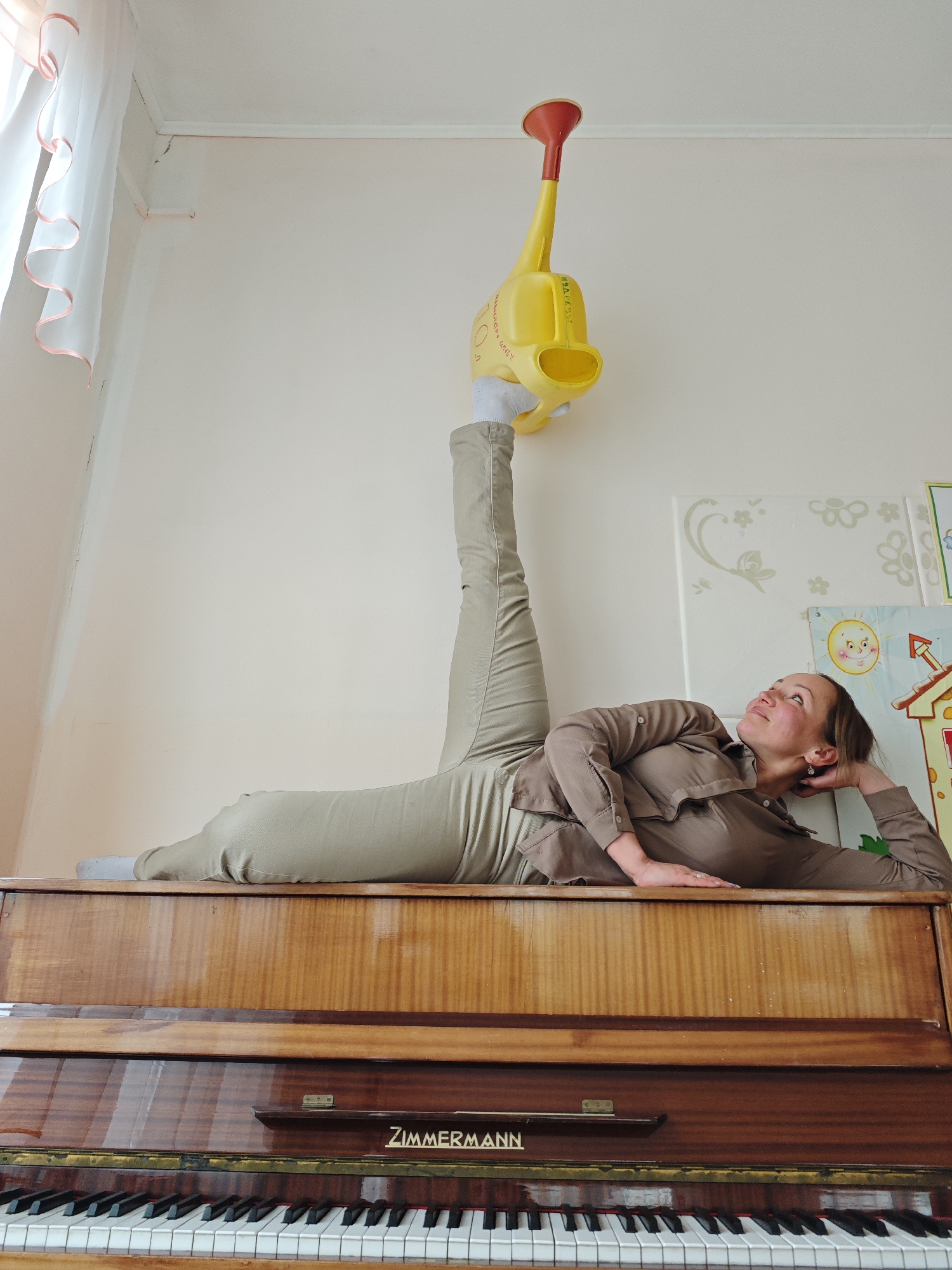 A woman is lying on top of a piano in a room, holding a yellow watering can with one leg extended upwards. The watering can is positioned so that it appears to be standing upright against the wall. The woman is wearing a brown button-down shirt and tan pants. The piano is dark wood with the name "Zimmermann" inscribed on it. Behind the woman is a wall with decorative drawings of flowers and a sun, possibly children's artwork. A window with a white curtain is visible in the upper left corner. The location is implied to be Markovskii, Russia based on the prompt instructions.