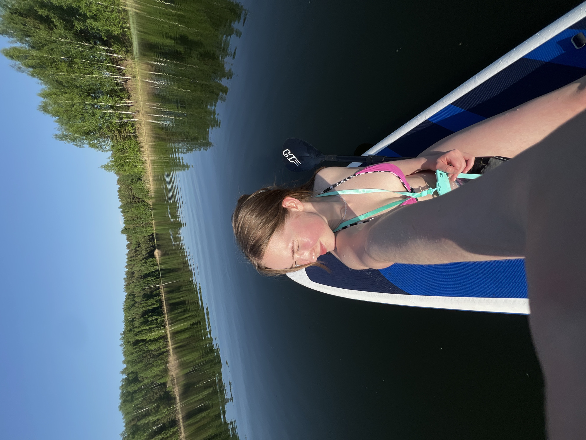 A woman is lying on a paddleboard in a body of water, possibly a lake or a wide river. She is wearing a pink and turquoise bikini. A paddle is lying next to her on the board. The surrounding environment includes a wooded area visible on the shoreline with a clear blue sky above. The water is dark and reflects the trees. The location is assumed to be near Novyi, Russia based on the implied tags.