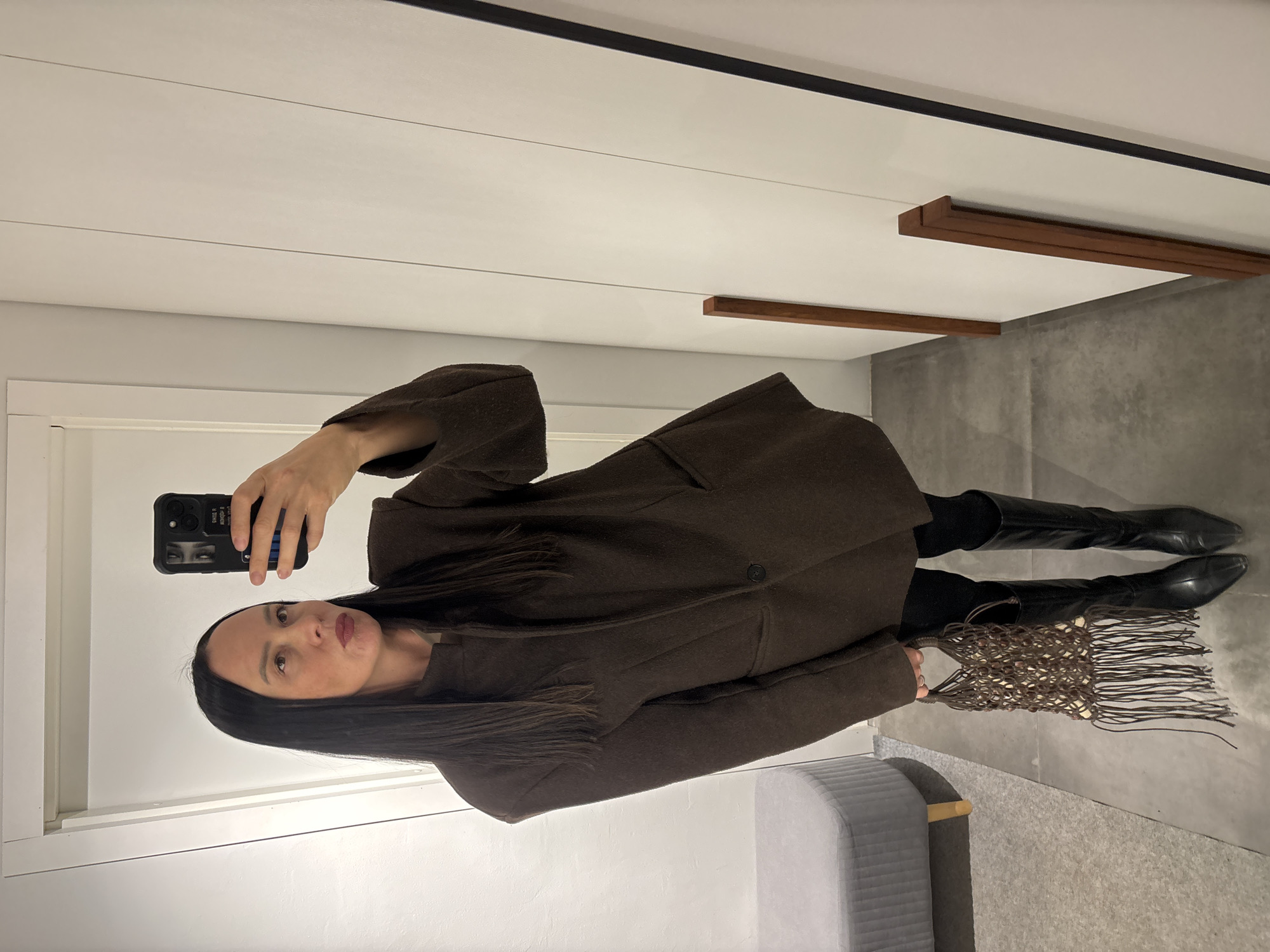 A woman is depicted taking a full-length mirror selfie indoors in Chaikovsky, Russia. She holds a black smartphone with an eye design and some text on its case in her right hand. The woman has long, dark hair and is wearing dark red lipstick, with a neutral facial expression.
She is dressed in a dark brown, possibly wool, coat or jacket, which features a single button and pockets. Underneath, she wears black long-sleeved clothing and black leggings or pants. Her footwear consists of black, knee-high boots with pointed toes. In her left hand, she carries a unique brown, fringed bag with a macrame-like texture.
The setting is a modern indoor space. The background shows light-colored walls and a dark grey concrete floor. Horizontal wooden strips are visible on the wall, adding a minimalist detail. A grey upholstered bench or ottoman is positioned in the lower left corner of the image. The lighting appears to be consistent with an indoor environment, without specific cues for time of day or weather.
Visible text on the smartphone case includes parts of words like "TRUE" or "TRUST" and other indistinct characters.