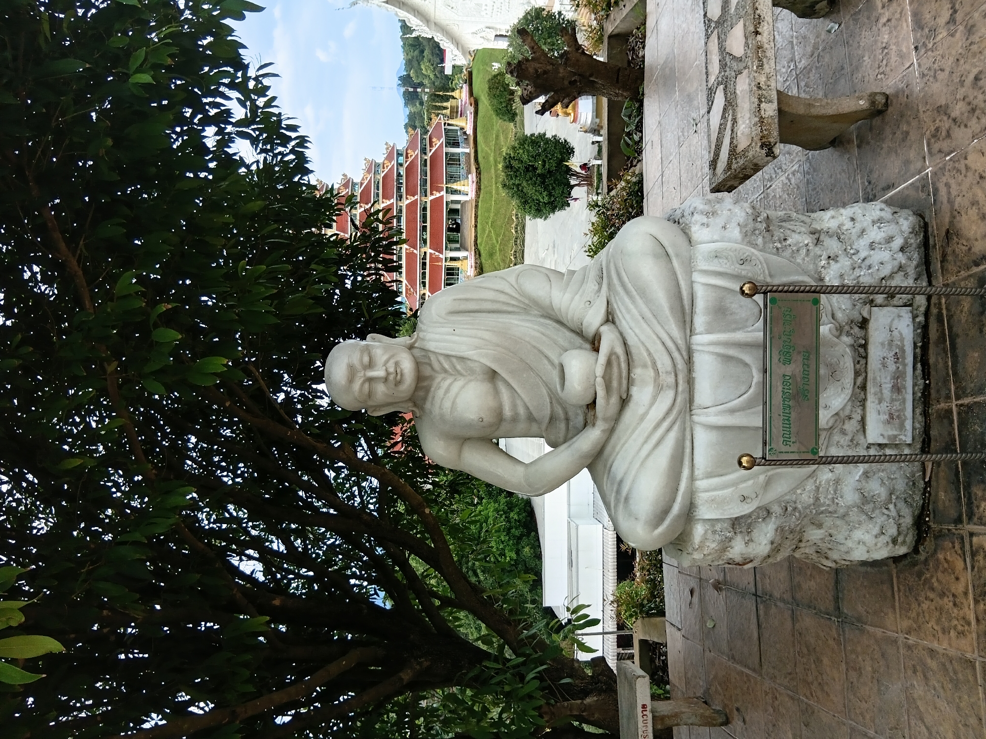 This image shows a white marble statue of a seated monk in a meditative pose. The statue is placed on a rough, white stone base. In front of the statue, a green plaque with Thai script is displayed on a metal stand. To the right of the statue, a weathered stone bench with a mosaic top is visible. The foreground is paved with square tiles. In the background, a large, multi-tiered building with a red roof and ornate gold details stands prominently. Green trees and landscaping surround the building. The sky above is partly cloudy. The overall scene appears to be an outdoor religious or cultural site, likely a temple or monastery, situated in a lush, green environment. The inscription on the plaque, when translated, refers to a revered monk, possibly "Luang Por Khun Parisuttho," a famous Buddhist monk from Northeastern Thailand. The presence of the temple structure suggests this location is in Thailand. Given the context of the statue and temple architecture, the location is identified as Ban Lao Phatthana (บ้านเหล่าพัฒนา), Thailand.
