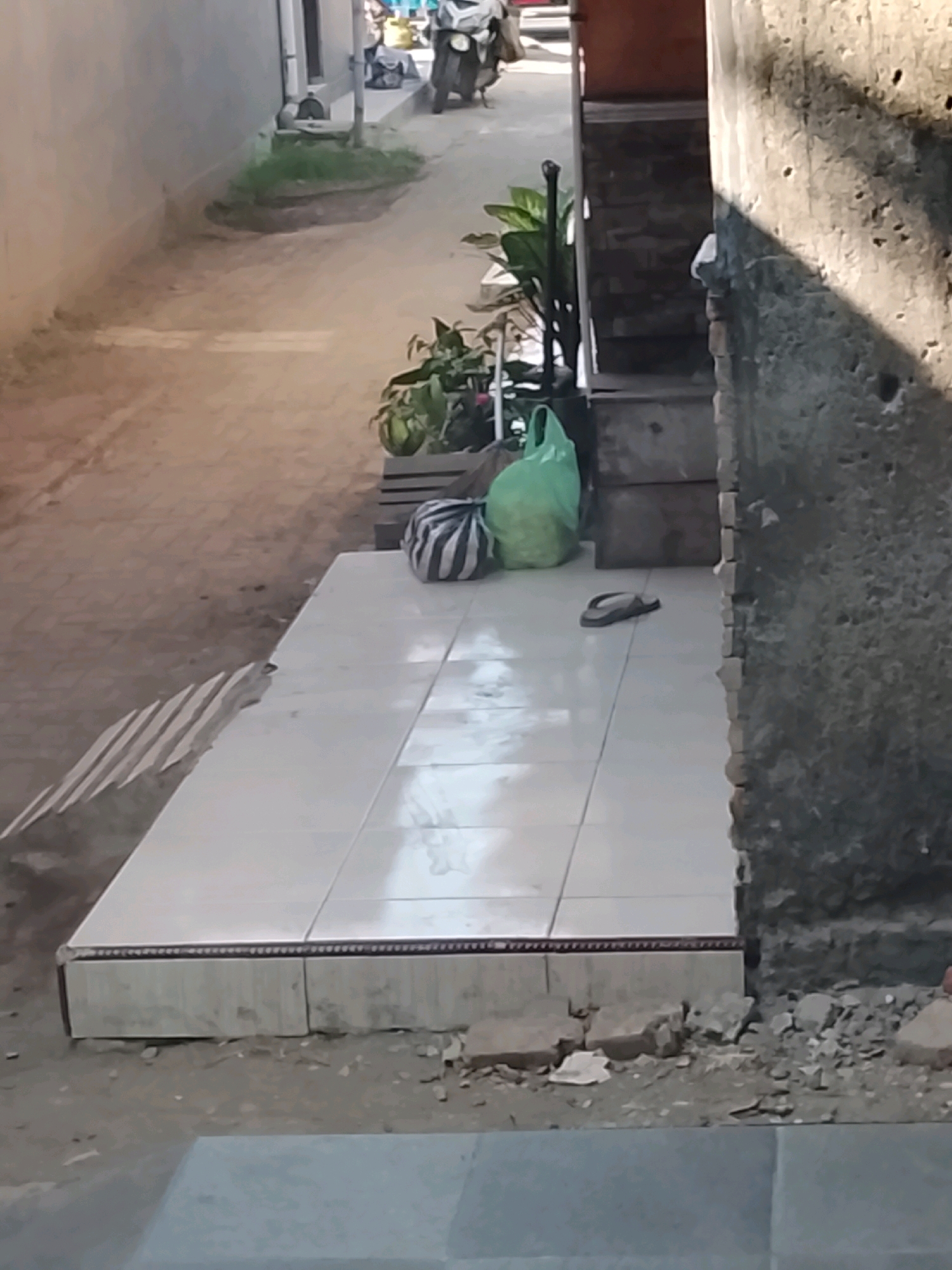 The image depicts an outdoor alleyway with a tiled walkway in the foreground. On the tiled surface, there is a single dark sandal. To the left of the sandal, there is a green plastic bag and a striped duffel bag. Further back, to the left of the tiled area, a pathway is visible with a patch of grass and some greenery. In the background, a brick wall and a set of stairs are partially visible. The overall scene suggests a residential or back alley setting in a warm climate, possibly in Ujungrusi, Indonesia, given the context. The lighting suggests it is daytime, with shadows cast by an unseen structure.