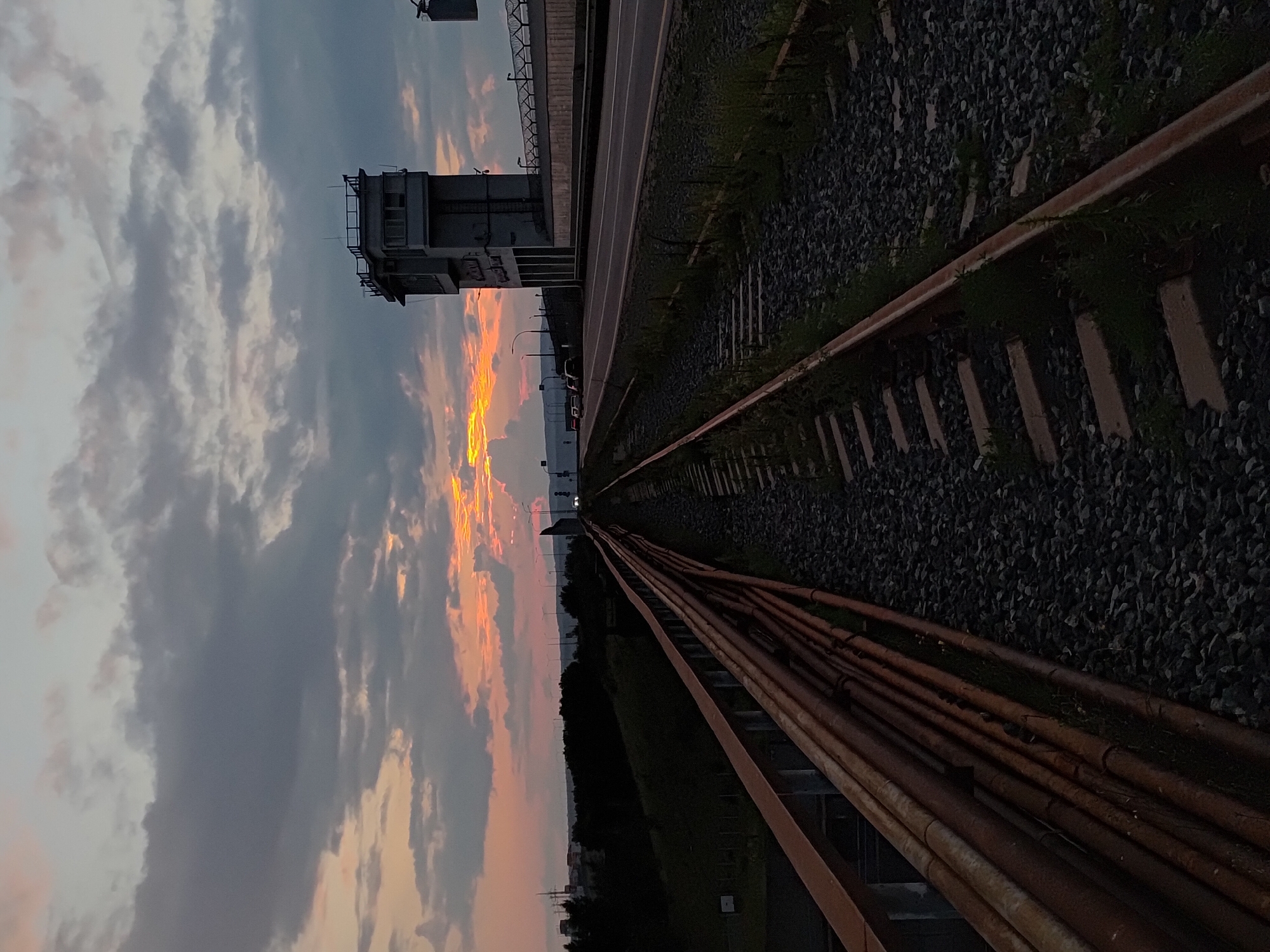 The image depicts a railway track in the foreground, running parallel to a road in the mid-ground. The sky is filled with dramatic clouds, with streaks of orange and yellow light suggesting a sunset. A tall, industrial-looking building with windows and a railing on top stands beside the road. Further in the distance, the silhouettes of trees and urban structures are visible against the dusky sky. The overall scene has an industrial and somewhat desolate feel, possibly located near a dam or a power plant given the presence of the building and its proximity to water, which is suggested by the landscape. It is likely an area within the city of Chaikovsky, Russia, known for its industrial infrastructure, particularly related to the Votkinsk Dam. There are no people visible in the image.