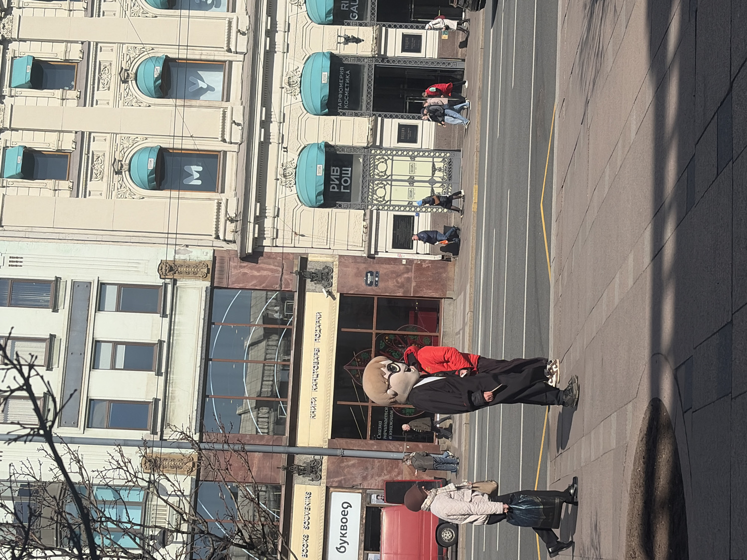 The image displays a lively street scene in Saint Petersburg, Russia, under clear, bright daylight, evidenced by strong shadows. Multi-story buildings with intricate architectural details line the street. Their ground floors host various shops, prominently featuring "РИВ ГОШ" (RIVE GOSH), a perfumery and cosmetics store with turquoise awnings, displaying the text "ПАРФЮМЕРИЯ КОСМЕТИКА" (PARFUMERY COSMETICS) and logos including an 'M' and a stylized bunny. Further along are "КАЙ КАНЦТОВАРЫ ПОДАРКИ" (KAI STATIONERY GIFTS) and "буквоед" (Bukvoed), a bookstore, indicated by "BOOKS SOUVENIRS" and the phrase "Сказка начинается" (The fairy tale begins).
On the sidewalk, various pedestrians are engaged in daily activity. The most notable subject is a person in a mascot costume, featuring a large, light-brown, round head with prominent eyes and a snout, resembling an anthropomorphic character. This figure is dressed in a dark suit jacket over a bright red top and dark trousers, standing near the street curb. Other pedestrians include a woman in a light-colored coat and a red beret, seen walking past a red vehicle, and several other individuals in the background. The street is an asphalt road with distinct white and yellow lane markings. The scene captures a typical urban environment on a sunny day.