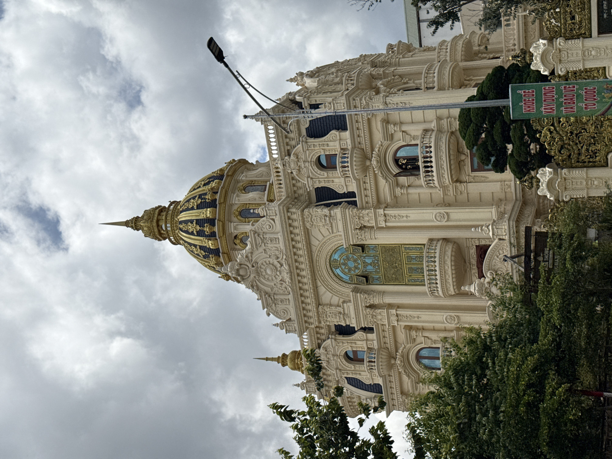 The image shows a building in Da Lat, Vietnam, characterized by an ornate architectural style with a golden dome. The sky is overcast with gray clouds. There is greenery surrounding the base of the building, and a sign is visible along the building’s side. A modern light post is visible near the dome. The architectural style of the building is predominantly European-inspired.
