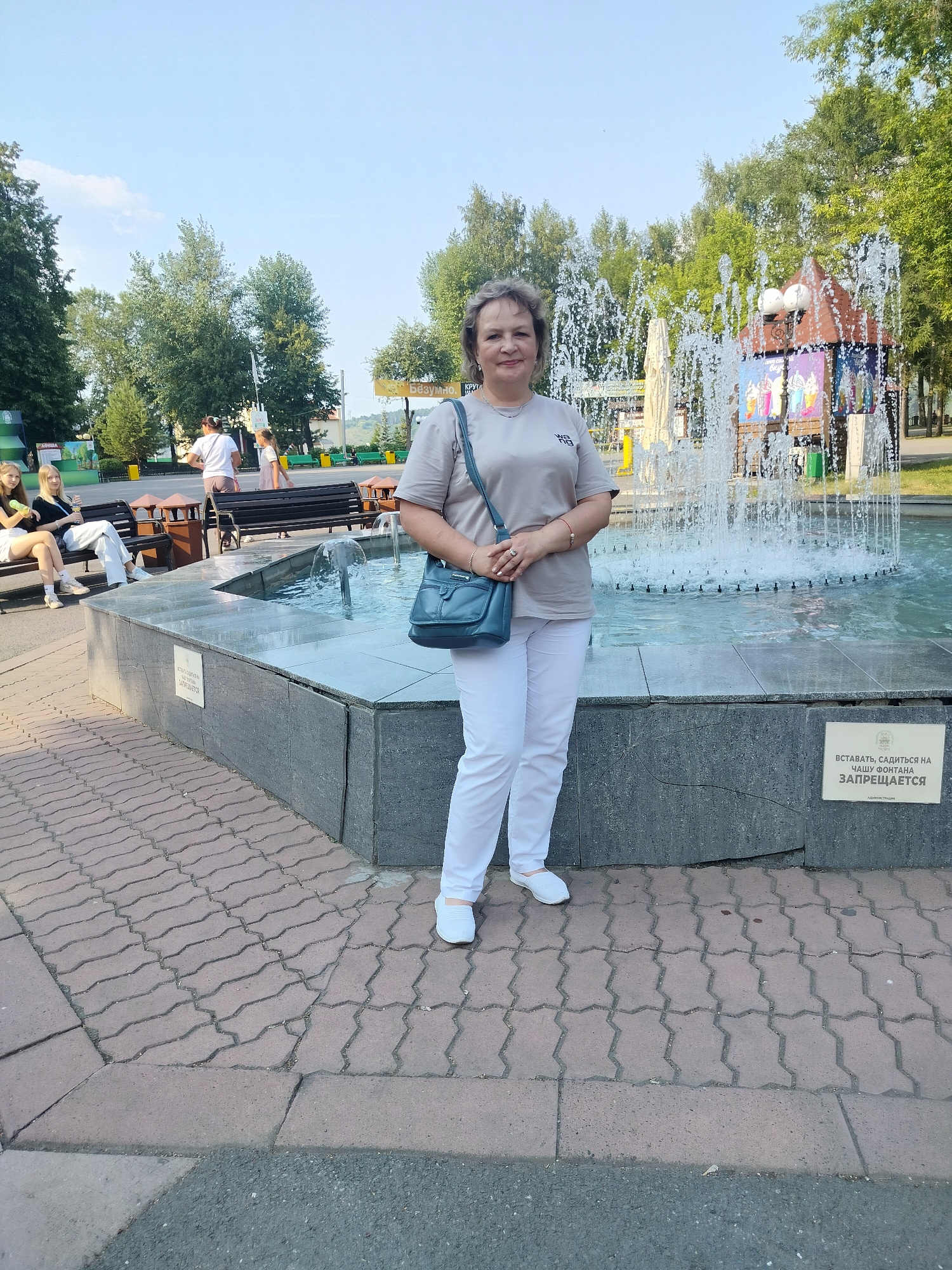 The image shows a woman standing in front of a fountain in the city of Kemerovo, Russia. The woman is wearing a beige t-shirt and white pants, and is carrying a blue purse. She is smiling and looking at the camera. The fountain is located in a park with trees and benches. There are other people in the background, some of whom are sitting on benches and others who are walking. The sky is blue and there are some clouds. The fountain is made of stone and has water spraying from it. There is a sign on the side of the fountain that says "ВСТАВАТЬ, САДИТЬСЯ НА ЧАШУ ФОНТАНА ЗАПРЕЩАЕТСЯ" which translates to "It is forbidden to stand or sit on the fountain bowl".