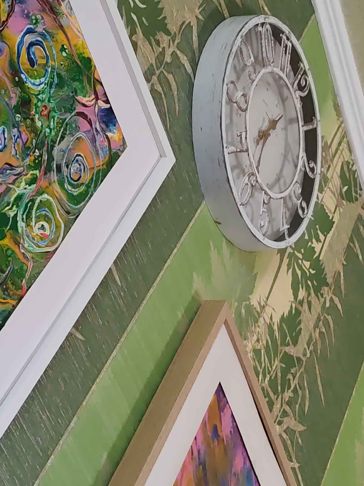 The image shows a close-up of a wall with a patterned wallpaper and two framed art pieces. A white framed abstract painting is visible on the left, and a second framed artwork with a light brown frame is in the lower right corner. A decorative white clock with a distressed finish and gold-colored hands is mounted on the wall between the two art pieces. The wallpaper has a green background with a pattern resembling bamboo or reeds in a lighter, almost gold color. The image is taken from a slightly elevated angle, looking down towards the right. There are no people visible in the image. The location is identified as Rostov-on-Don, Russia.
