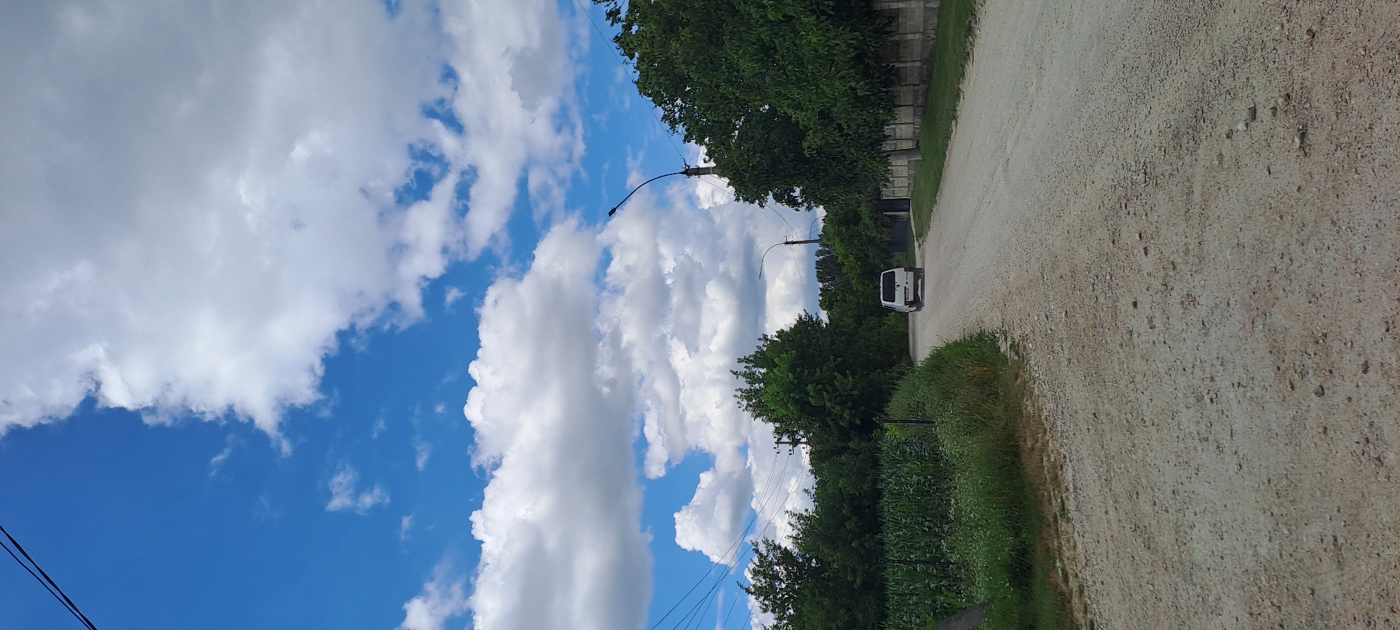 The image captures a dirt road in Rădoaia, Moldova, under a bright blue sky dotted with fluffy white clouds. On the left side of the road, a field of green corn stalks stands tall, bordered by a sparse fence and overgrown grass. The road itself is unpaved and appears to be made of gravel and dirt. A white minivan is visible in the distance, driving away from the camera towards the right side of the frame. The right side of the road features a low concrete fence and a line of lush green trees. A solitary lamppost stands on the right side of the road, its pole tall and curved. Overhead, power lines are strung across the sky. The overall impression is a rural, somewhat undeveloped setting in the daytime.