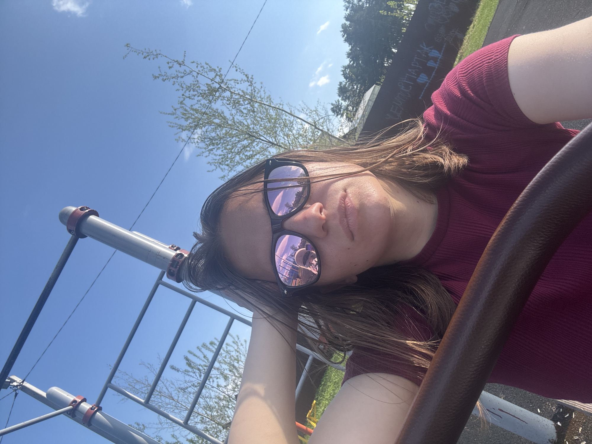 A selfie photograph taken in a park on a sunny day in Раздолье, Russia. A woman wearing sunglasses and a maroon ribbed shirt is the focal point. The sunglasses reflect a playground scene. To the left, a metal playground structure with horizontal bars is visible against a bright blue sky with scattered clouds. In the background, a wall with graffiti, grass, and trees can be seen.