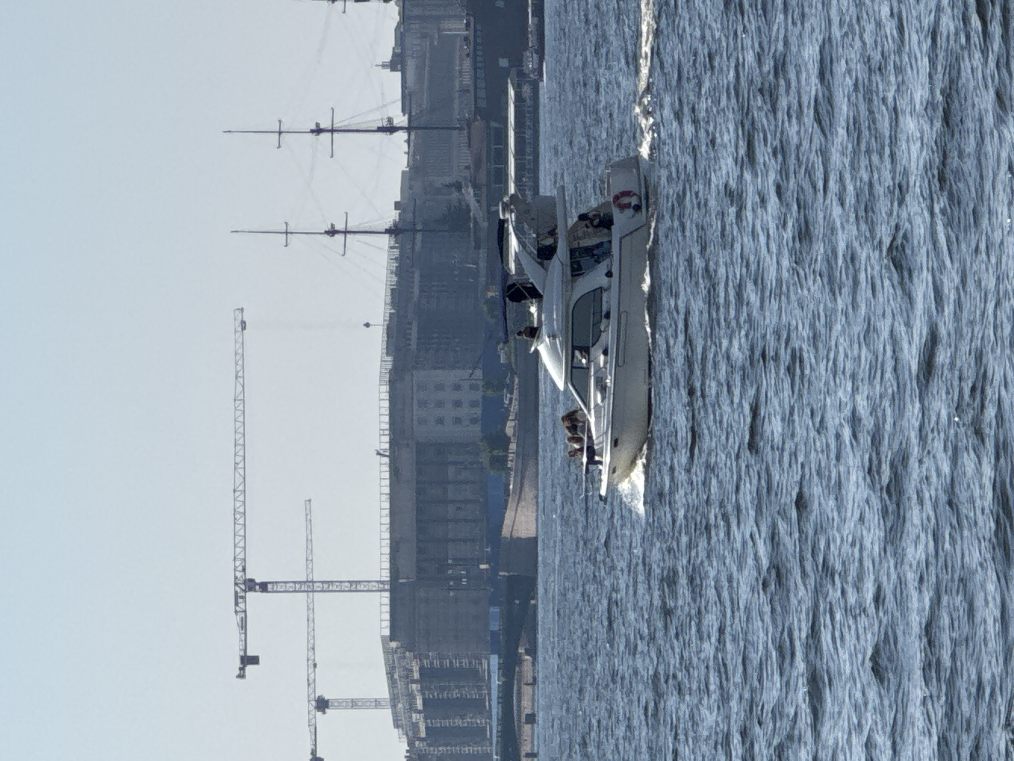 The image shows a cityscape with a body of water in the foreground. A white motorboat is moving through the water, creating a wake. There are several people visible on the boat. In the background, there is a large construction site with cranes, and further back, city buildings and what appear to be masts of old sailing ships. The sky is a pale, hazy blue, suggesting it is daytime, possibly overcast or early morning/late afternoon. The water appears choppy. The overall scene depicts a busy urban waterfront, possibly undergoing development, with recreational activity on the water. Given the mention of Saint Petersburg, Russia, it is likely the Neva River or a canal.