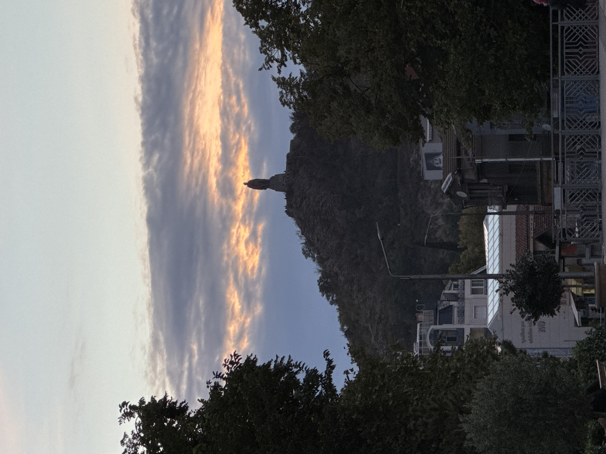 The image shows a view towards the Alyosha monument on Bunardzhik Hill in Plovdiv, Bulgaria. The sky is partly cloudy with the sun setting, casting orange and pink hues on the clouds. The monument stands on top of the hill, silhouetted against the sky. In the foreground, there are trees, buildings, and a street lamp. The overall scene suggests an evening in the city, with a mix of natural beauty and urban landscape.