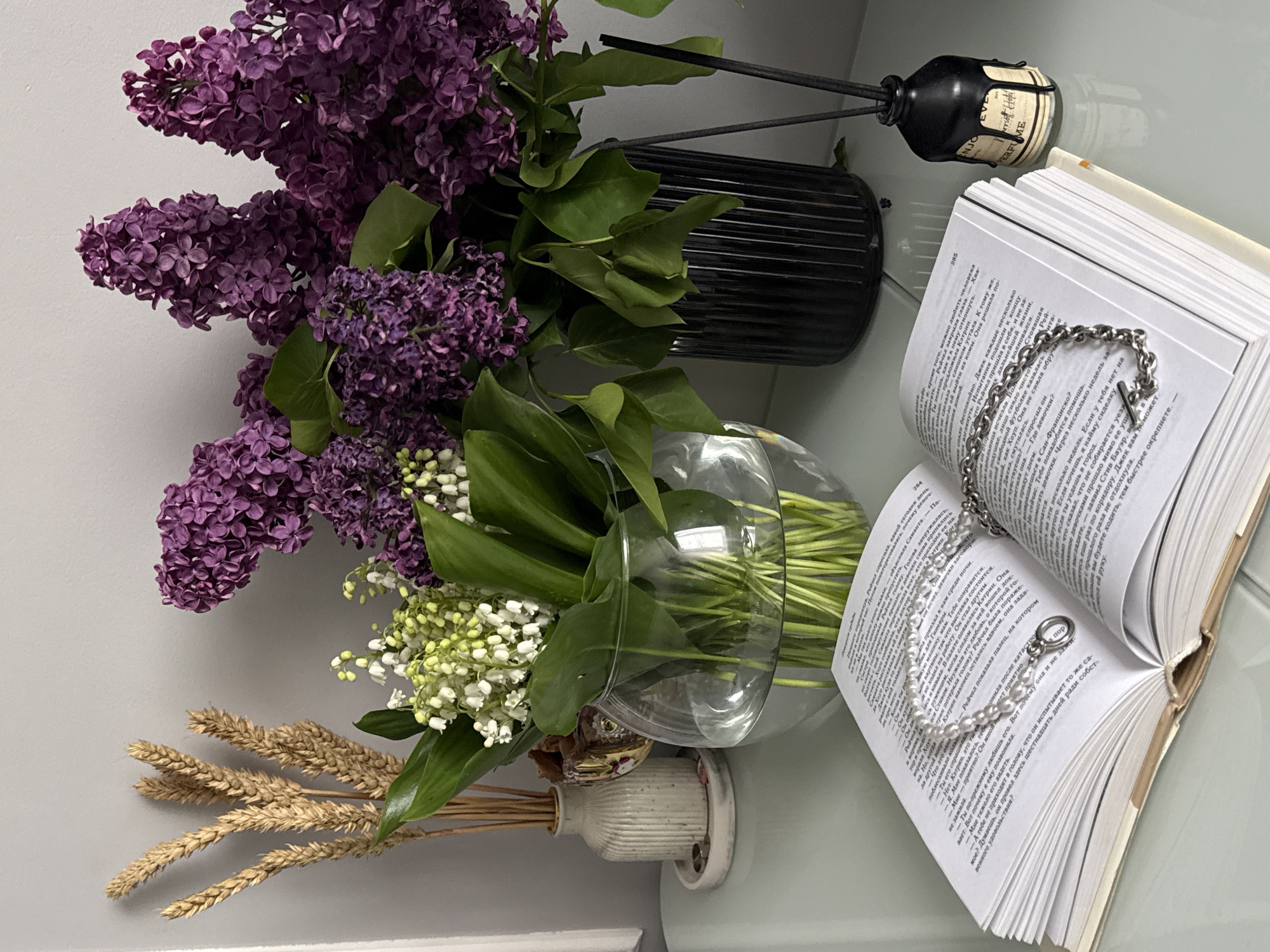 The image, captured from an overhead perspective, displays a still life arrangement of various objects on a light-colored surface, with the entire composition rotated 90 degrees clockwise from its natural orientation.
On the left, a lush bouquet of vibrant purple lilacs with green leaves fills a tall, black, ribbed vase. Adjacent to it, a clear glass bowl holds delicate white lily of the valley flowers alongside green foliage. Below these flowers, several stalks of dried, golden wheat are arranged in a small, vertically ribbed white ceramic vase.
To the right, an open book with numerous pages lies flat, revealing text written in a Cyrillic alphabet. Page numbers "285" and "284" are visible at the top corners of the open pages. A silver chain bracelet with a T-bar clasp rests on the left page, while a white pearl bracelet with a circular clasp is laid across the right page. Behind the book, a black diffuser bottle containing dark reeds is visible. A label on the bottle is partially legible, showing the words "ENJOY PERFUME".
The setting appears to be indoors, likely a table or shelf, with a plain light-colored wall forming the background. The natural lighting suggests it is daytime. The overall arrangement creates an atmosphere of tranquility and aesthetic pleasure.