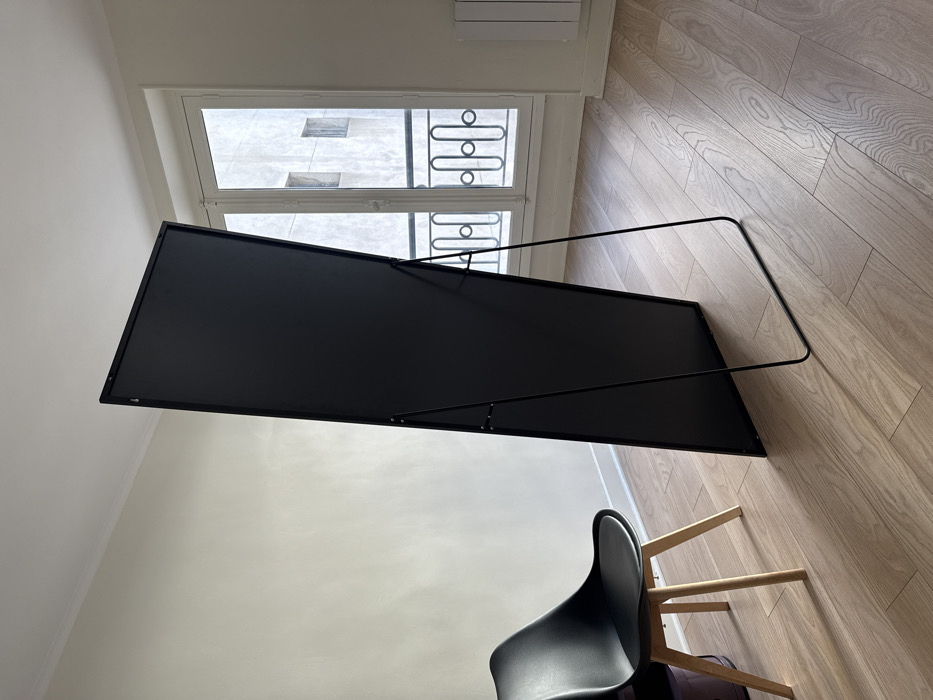 The image depicts an interior room, characteristic of a Parisian apartment in Paris, France. The room features light cream-colored walls, white baseboards, and a light-toned wooden floor laid in planks. A tall, rectangular full-length mirror, displaying its matte black rear surface and supported by a black metal kickstand, stands angled towards the left on the right side of the frame. To the left, a modern chair with a black molded plastic seat and backrest, supported by four light-colored wooden legs, is positioned. Partially obscured behind the chair's left leg is a dark, cylindrical object. In the background, a large white French window, composed of multiple panes, offers a view to the exterior. An ornate black wrought-iron balcony railing is visible through the lower section of the window. Beyond the balcony, the facade of a light-colored building with multiple rectangular windows is visible. A white horizontal radiator is mounted on the wall beneath the window on the far right. No individuals are present in the scene.