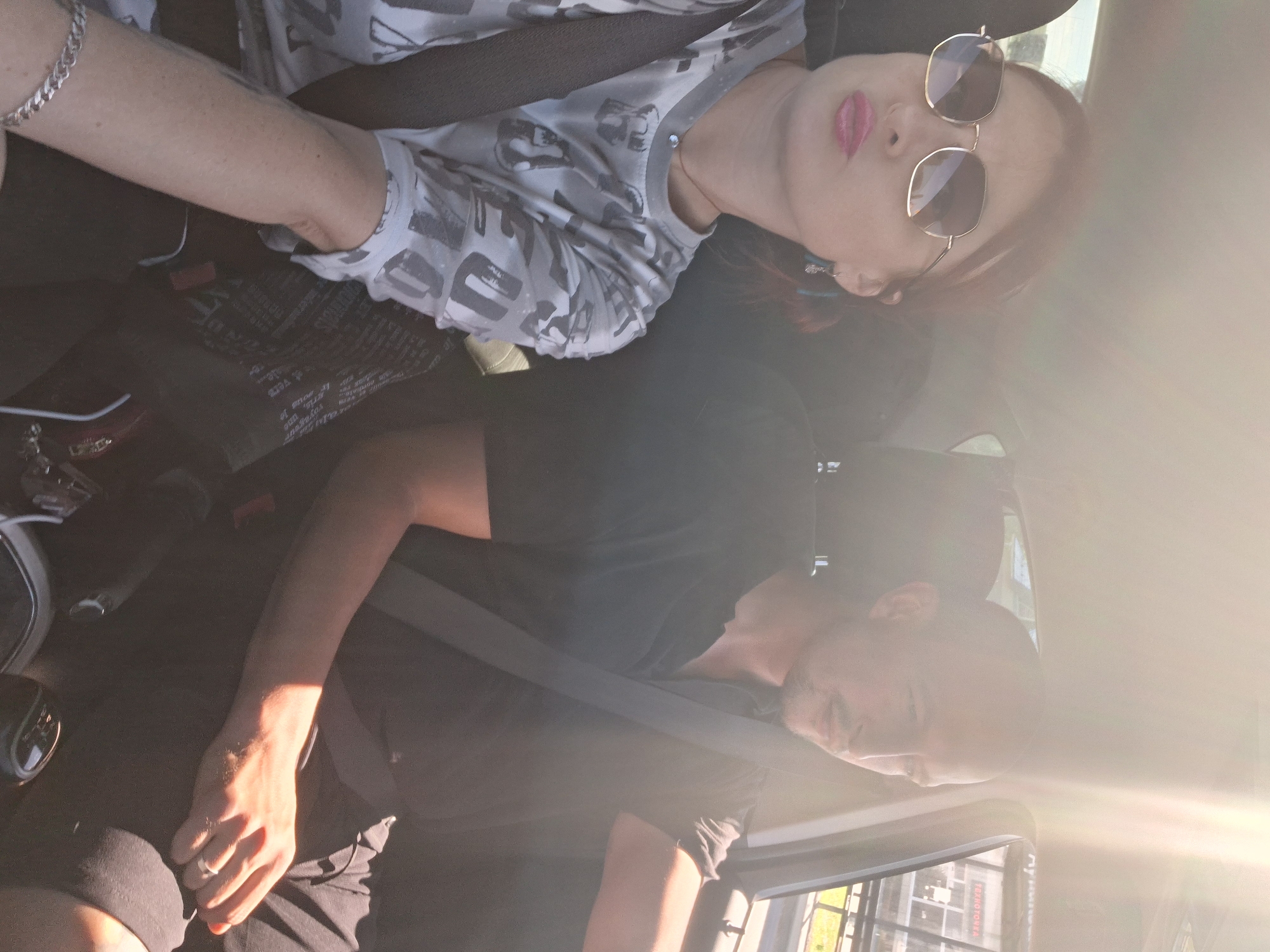 Two individuals, a woman and a man, are seated in the front passenger and driver seats of a car, respectively. The woman, on the left, wears sunglasses, a gray and white patterned shirt, and a necklace. She has a silver bracelet on her left wrist and is looking forward with pink lipstick visible. The man, on the right, is wearing a black polo shirt and appears to be driving. Both are wearing seatbelts. The interior of the car is visible, including the gear shift. Bright sunlight streams in from the right side of the car, creating lens flare and highlighting the right side of the man's face and arm. Outside the car, through the windows, a blurred cityscape is visible, with buildings and what appears to be signage suggesting an urban environment. The presence of Cyrillic script on some of the signs and the name "Myadzyel, Belarus" in the prompt indicates this scene is likely set in that location.