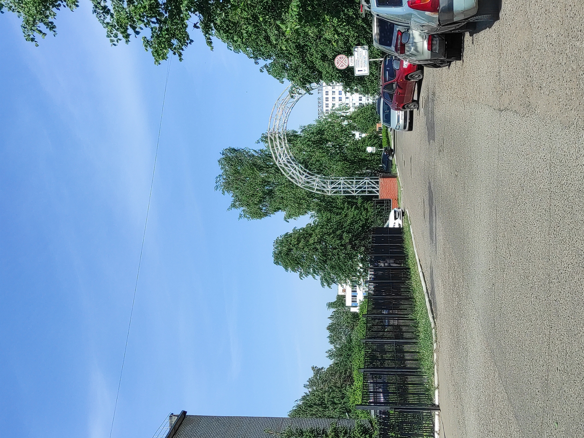 The image shows a street in the city of Chaikovsky, Russia. The street is paved and lined with a black metal fence on one side. A decorative metal arch stands over the street. Several cars are parked along the curb. Trees are visible on both sides of the street and in the background, partly obscuring a white building. The sky is blue with some thin clouds.