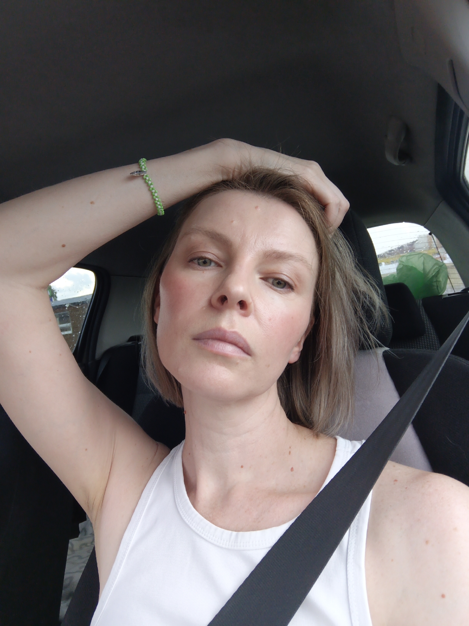 A medium shot captures a woman from the shoulders up, seated in the passenger seat of a car. She has fair skin, light brown hair, and is wearing a white tank top and a green and white beaded bracelet on her left wrist. Her left arm is raised, with her hand resting on top of her head. The background shows the interior of a car, including the headrest and seatbelt, and a blurred view of the exterior through the window, suggesting an urban or suburban setting. The woman appears to be looking directly at the camera with a neutral expression. The image was taken in Chaikovsky, Russia.