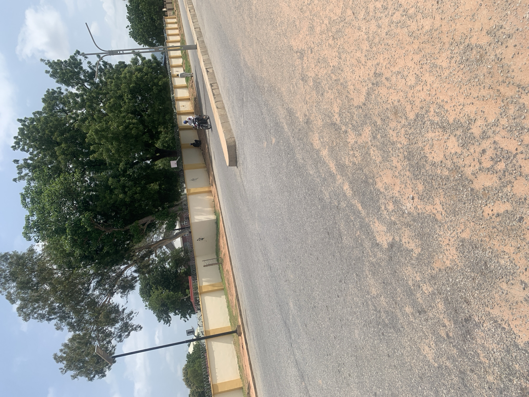 The image shows a street scene in Jalingo, Nigeria. A road with a sandy shoulder occupies the foreground. In the background, a beige wall topped with barbed wire runs along the street. Behind the wall, there are several large trees with dense green foliage and some buildings are visible. Two people are on a motorcycle, with one driving and the other as a passenger, traveling on the road. Another person is walking further down the road. A street lamp is visible on the left side of the image. The sky is partly cloudy.