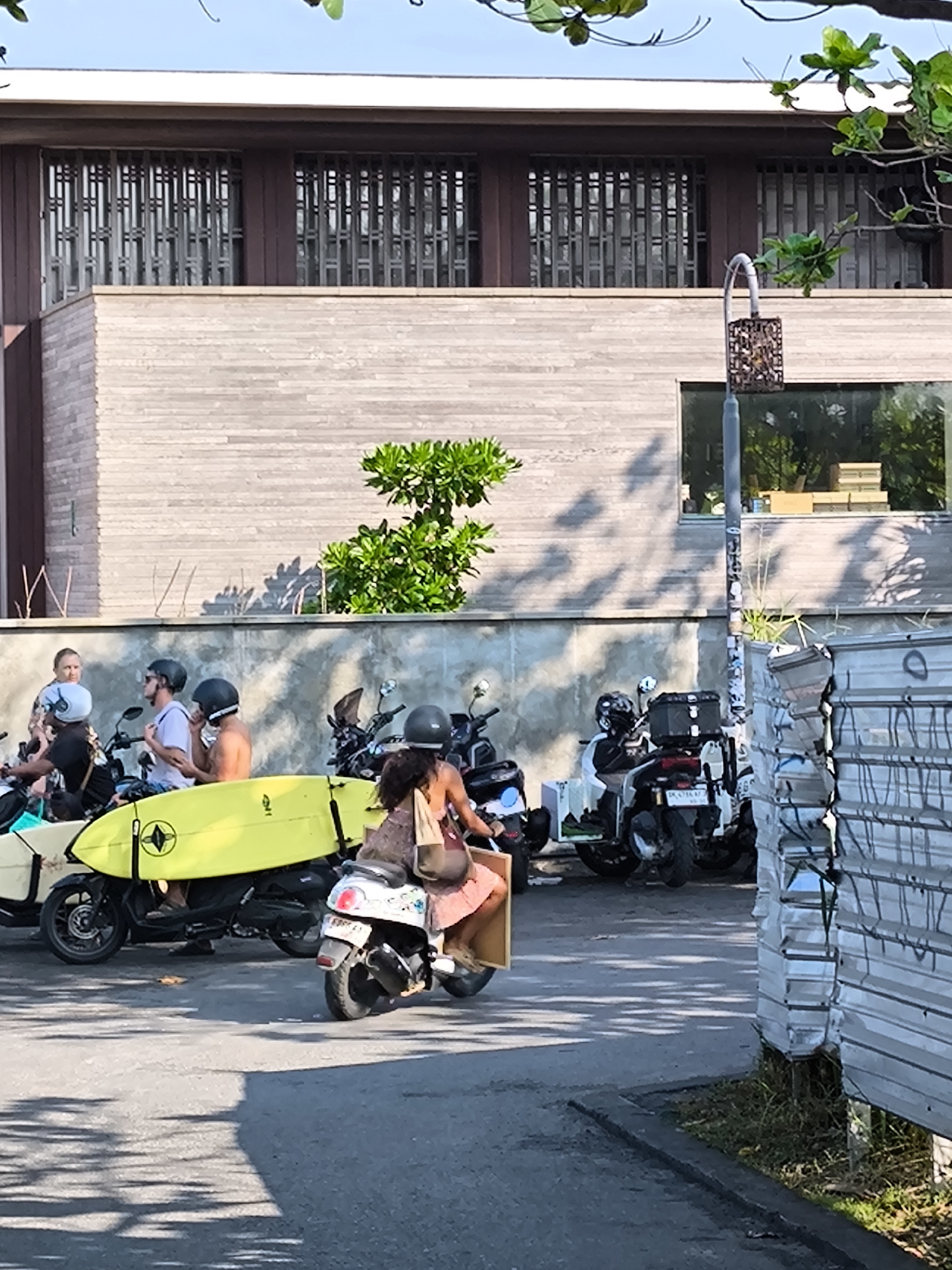 The image depicts a street scene in Canggu, Indonesia. In the foreground, a woman wearing a helmet and light-colored dress rides a white scooter away from the camera. To her left, several parked scooters are visible, one carrying a bright yellow surfboard. These scooters seem to belong to a group of people, some shirtless or in light clothing, standing or sitting on their bikes. They seem to be socializing.
Behind the parked scooters and people, a modern building stands. It features horizontally-laid wooden slats, dark brown window frames, and decorative bars on the windows. A small tree or bush grows near the base of the building, just above a low concrete wall. To the right of the building, there is a street lamp.
In the extreme foreground, a stack of corrugated metal sheets stands against the curb, partly obscuring the scene. The lighting suggests it's daytime with strong sunlight creating shadows on the road.