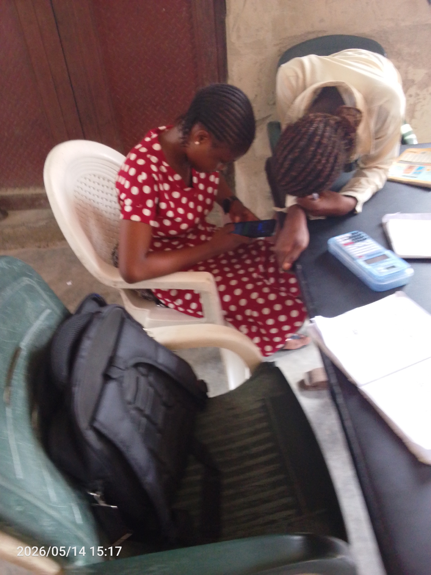 Two women are seated at a dark table in Lokoja, Nigeria. One woman is wearing a red polka-dot dress and is looking at a cell phone in her hands. The other woman is wearing a white shirt and is leaning over the table, her head resting on her arms. A blue calculator, papers, and notebooks are on the table. A black backpack sits on a green chair in the foreground. The location appears to be indoors, possibly a study or work area.