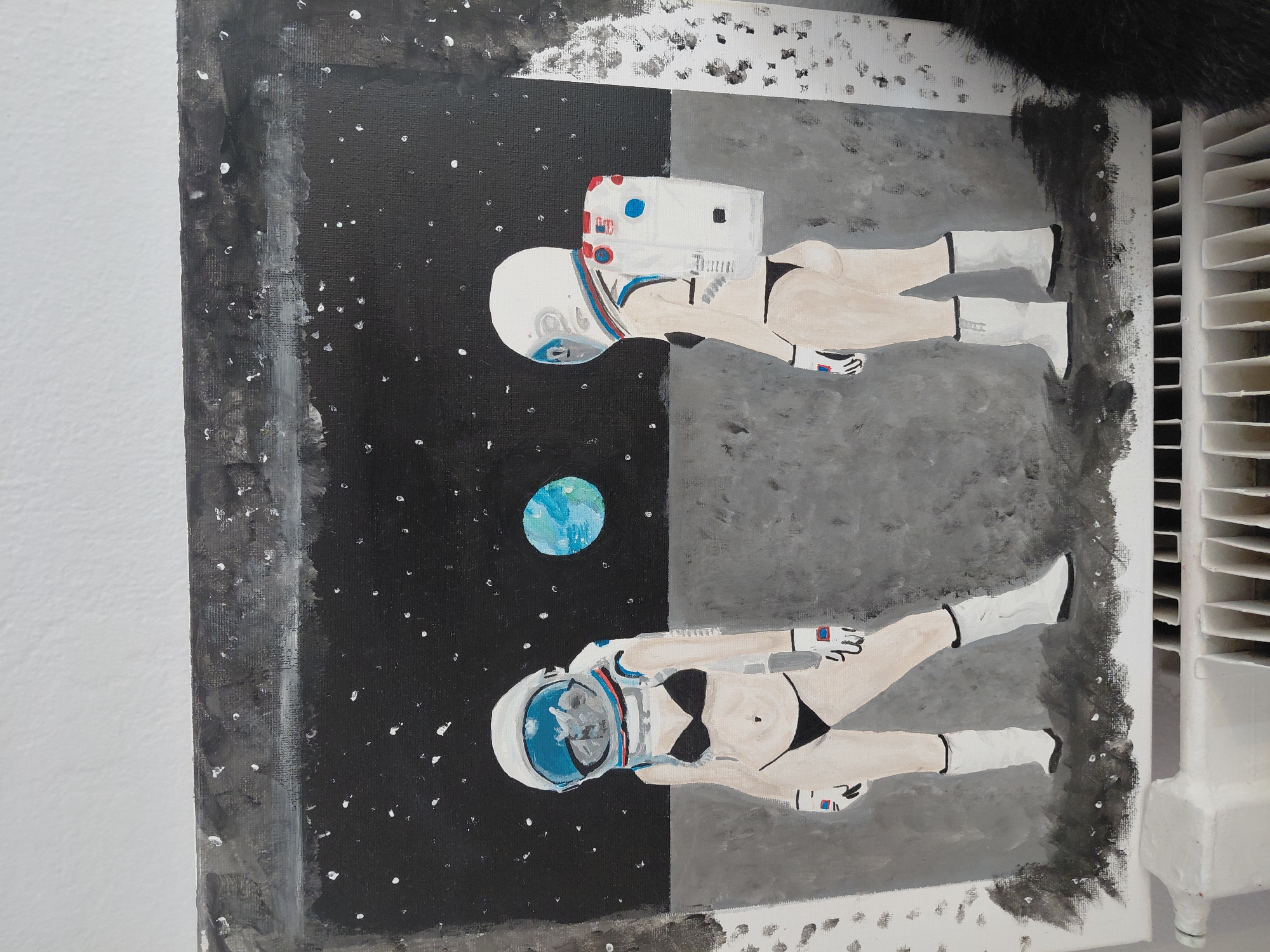 The image is a painting depicting two figures in astronaut suits, with their lower bodies exposed in bikinis, standing on a grey, rocky surface under a black, starry sky. A blue and green planet is visible in the distance. The painting is partially visible, with the bottom edge resting on a white radiator. The location is not specified within the image itself.