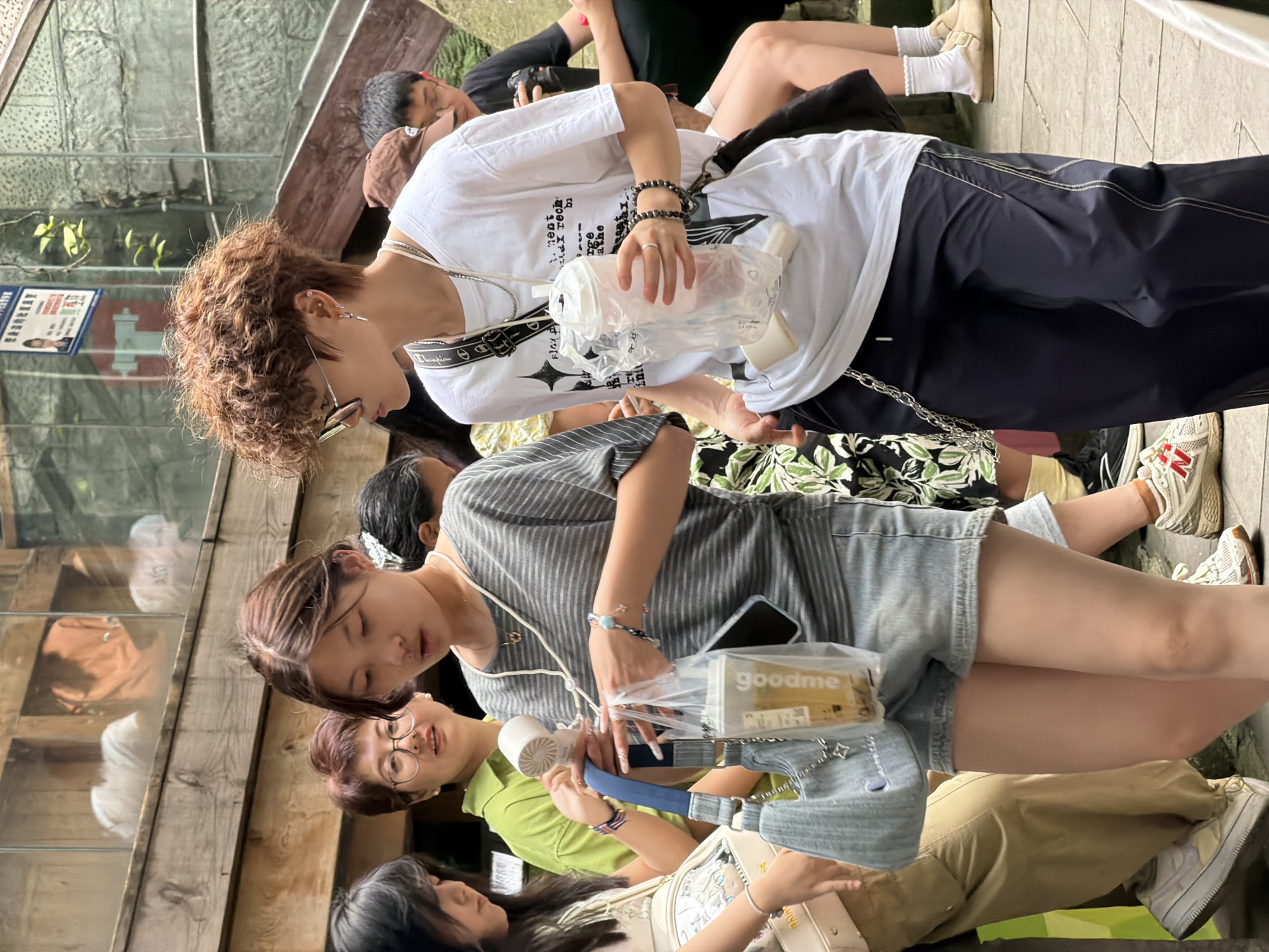 In Yuzhong District, China, a group of young adults are walking on a paved area adjacent to a stone structure with stairs. In the foreground, a young woman with short, curly brown hair and sunglasses is wearing a white t-shirt and dark pants. She is holding a plastic cup with a white lid and a straw. To her left, a young woman with long dark hair wears a grey striped t-shirt and denim shorts, carrying a light blue denim bag and a plastic bag containing a drink. Behind them, another young woman in a green shirt is visible, and further in the background, other individuals are seen, including a person wearing glasses and a dark t-shirt. The environment appears to be outdoors with wooden structures and greenery.