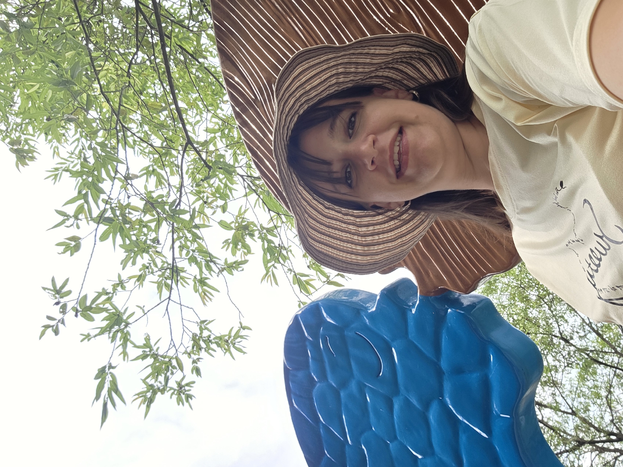 A low-angle, wide shot shows a woman wearing a wide-brimmed, striped sun hat and a light yellow t-shirt with a graphic design. She is smiling, with her head tilted slightly back, looking at the camera. To her left is a large, bright blue sculpture with a textured surface resembling scales or faceted jewels. To her right, behind her head, is a large, curved, ribbed structure made of a brown material, possibly wood or a similar composite. The background consists of a bright sky with some clouds and lush green tree branches with leaves. The overall impression is of a selfie taken outdoors, likely in a park or art installation, with a playful and candid atmosphere. The context of Gapcheon-myeon, South Korea is not visually apparent from the image itself.