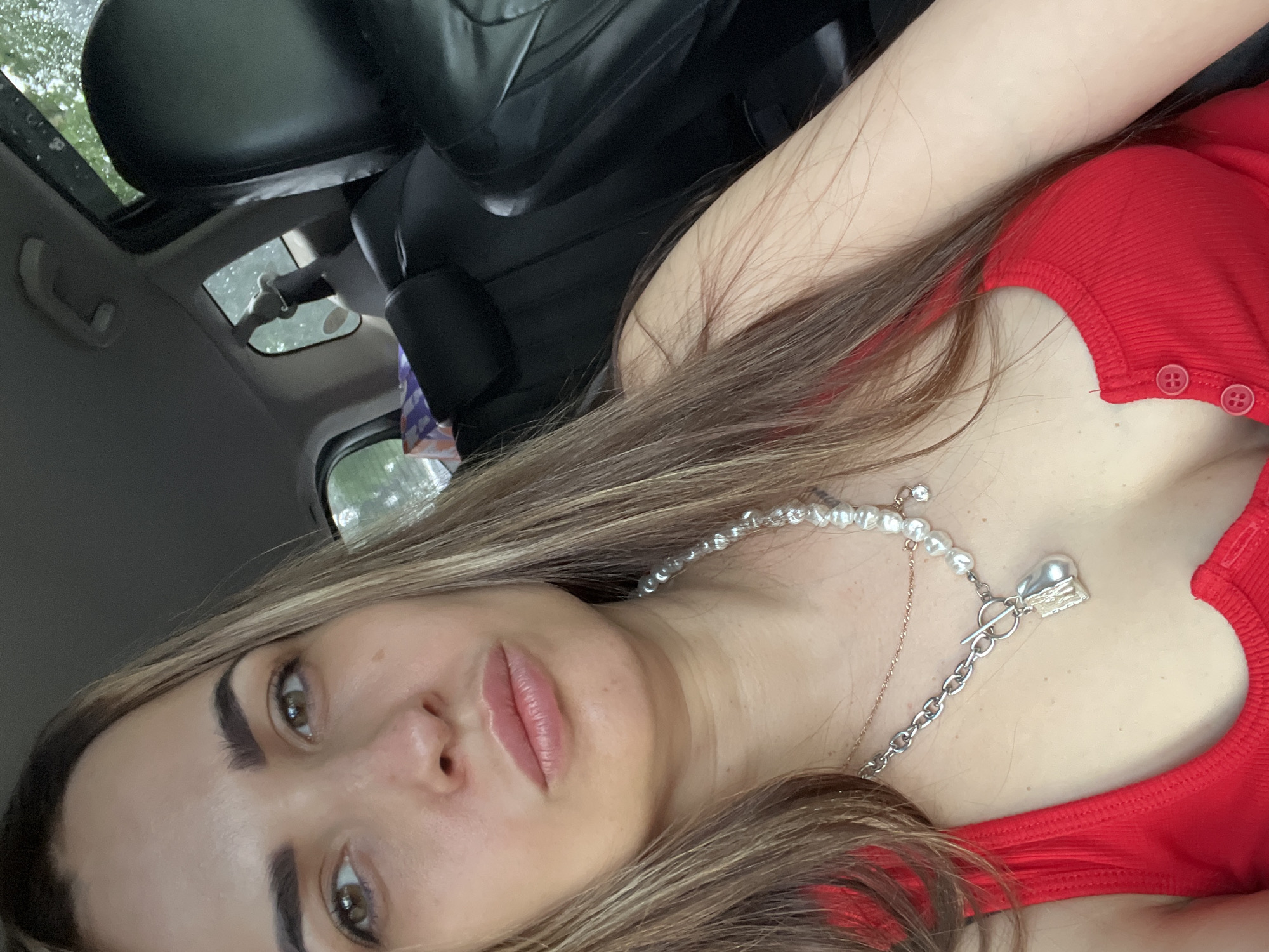 A close-up, tilted portrait captures a young woman inside a car. Her face, chest, and right shoulder are prominently featured. She has light brown hair with subtle highlights, which cascades over her shoulder and chest. Her eyes, a warm brown, are directed towards the viewer, and she has a neutral expression with slightly parted, natural-looking lips. Her eyebrows are well-defined.
She is wearing a ribbed red top, possibly a tank top or camisole, with two small red buttons visible on the strap or neckline on the right side of the frame. Around her neck, she wears two layered necklaces. The more prominent one is made of small white pearls, featuring a silver toggle clasp that connects to a thicker silver chain. This chain has a tear-drop shaped, possibly pearl, pendant. A delicate, thinner gold-colored chain is worn underneath the pearl necklace.
The background reveals the interior of a car. A black leather car seat is visible behind her, and above, a sunroof shows glimpses of the outside, which appears to be overcast or raining, with visible droplets on the glass. The car's interior ceiling is light grey. The lighting suggests it is daytime. There is no clearly readable text visible in the image.