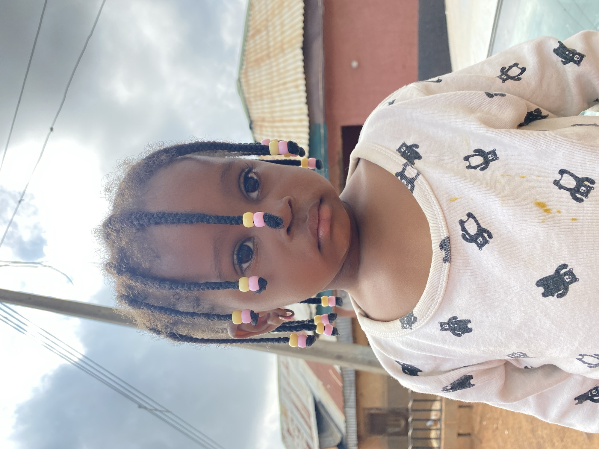 A young girl with braided hair adorned with pink and yellow beads stands outside in Bukuru, Nigeria. She is wearing a white long-sleeved shirt with a pattern of black bears. The background shows a building with a corrugated metal roof and some overhead power lines. The lighting suggests it is daytime, with some cloud cover.