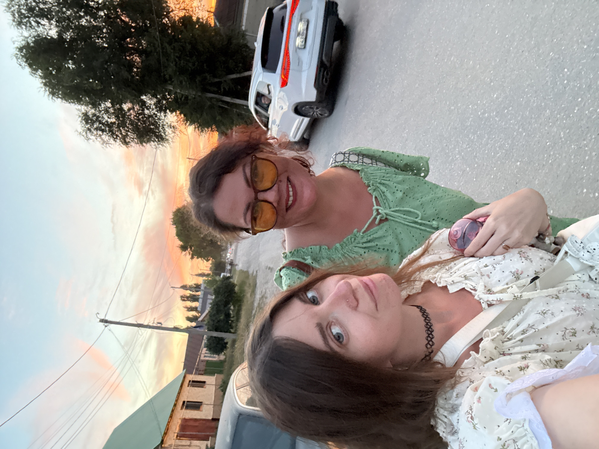Two women are taking a selfie on a street in Sol-Iletsk, Russia, during sunset. The woman in the foreground is wearing a white floral dress and a black choker necklace. The woman behind her is wearing a green dress and orange sunglasses. In the background, there are houses, trees, a car, and a power line. The sky is filled with orange and pink clouds.