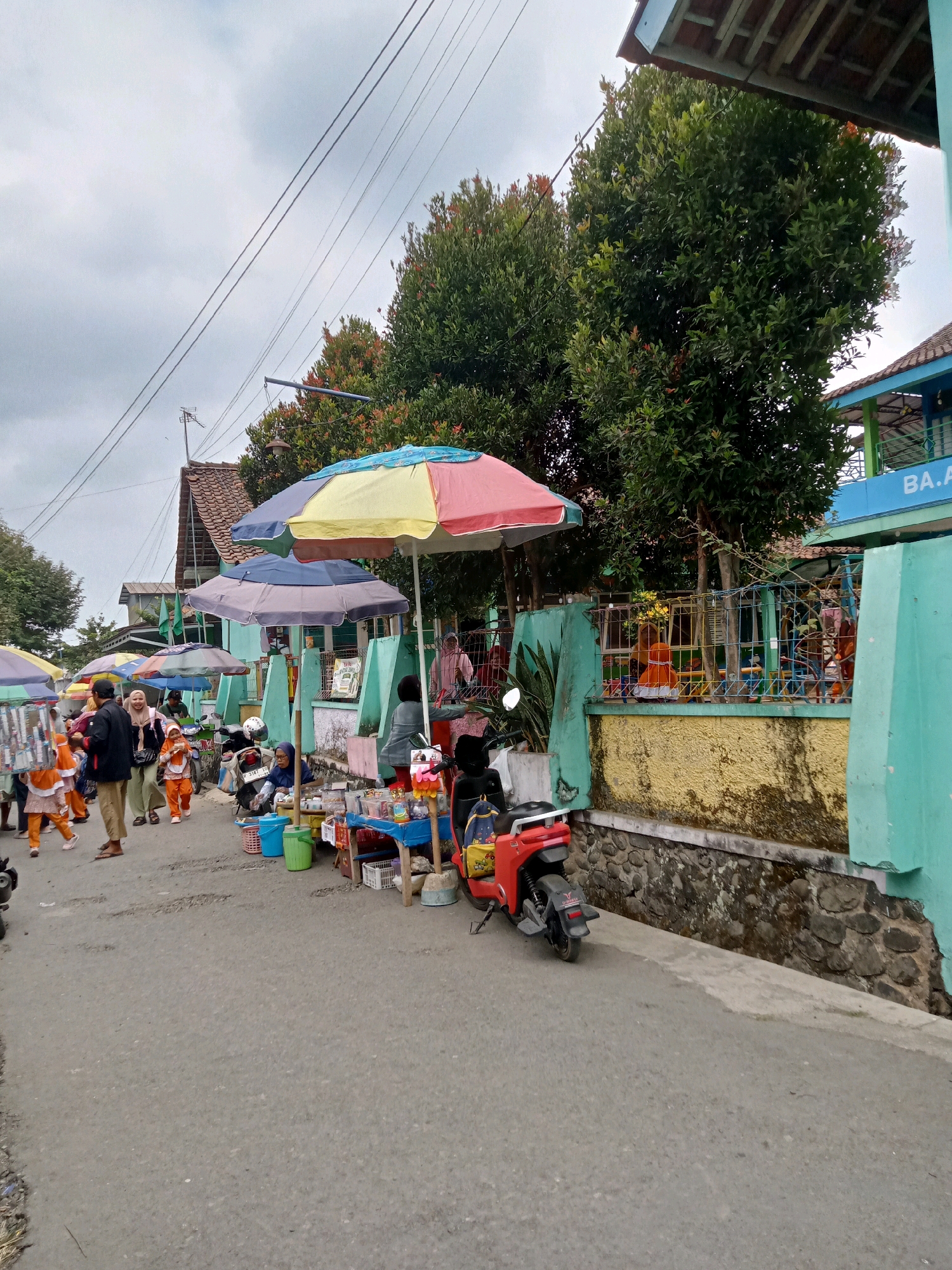 The image depicts a street scene in Gumiwang, Indonesia, with a variety of elements suggesting a lively marketplace or gathering. In the foreground, the asphalt road shows signs of wear and tear. To the right, a bright turquoise wall with a yellow textured lower section and a stone foundation borders the street. The wall is topped with a green metal fence, behind which a playground area with colorful equipment is visible. A sign on the turquoise building reads "BA.A".
Several stalls are set up along the street, indicated by colorful umbrellas providing shade. One stall on the right displays various goods, including what appear to be snacks or small items arranged on blue and red tables. A red electric scooter is parked in front of this stall.
On the left side of the street, more stalls are visible under umbrellas, one displaying what looks like small containers. A group of people are walking along the street, many of them children wearing orange outfits, possibly school uniforms or attire for an event. Adults are also present, some seemingly interacting with the stalls or other individuals. The background features trees with dense green foliage and reddish new growth, as well as buildings with terracotta roofs and turquoise accents. The sky is overcast with a cloudy, gray appearance. Overhead, electrical wires are strung across the scene, supported by poles.