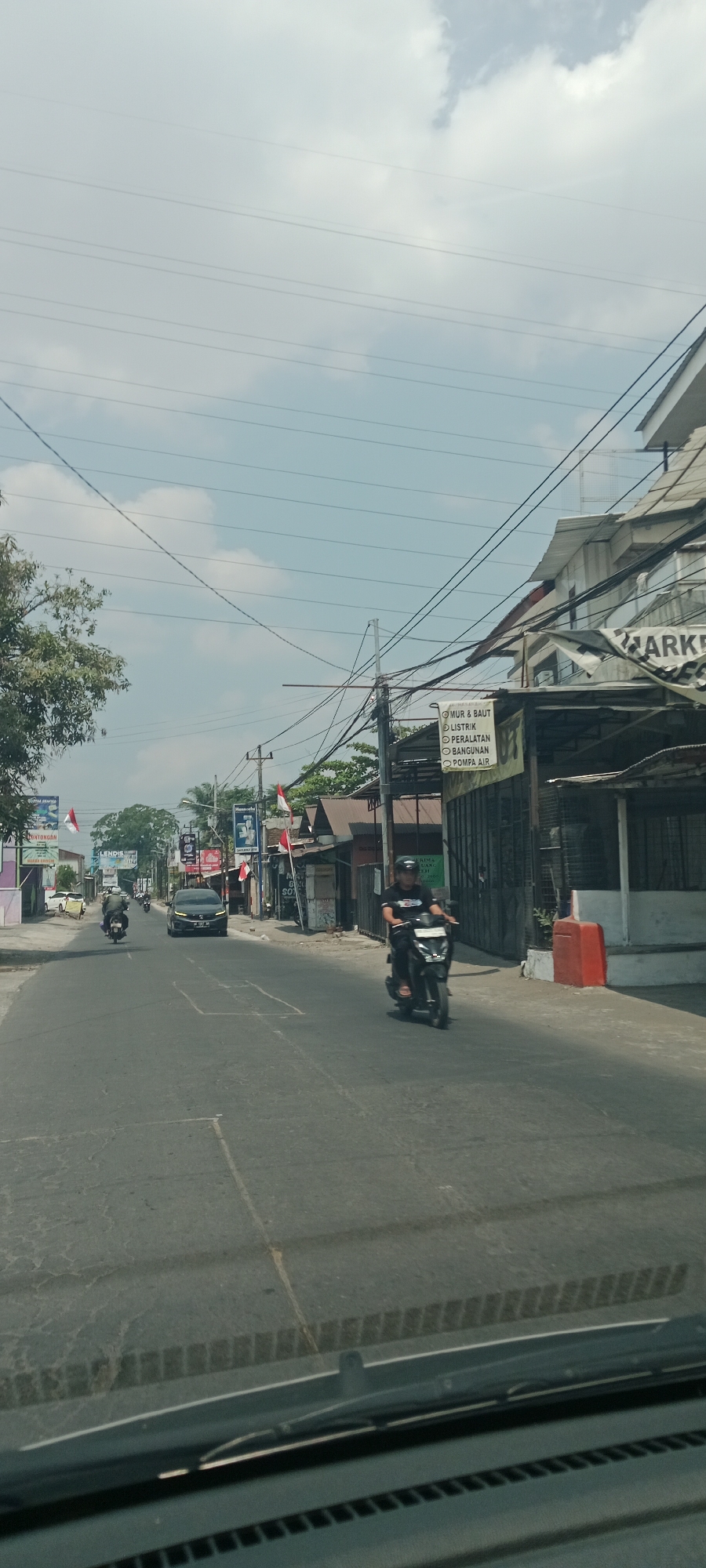 The image shows a street scene in Trihanggo, Indonesia. The perspective is from inside a vehicle, looking out at a paved road with some cracked sections and painted markings. On the right side of the road, there is a building with a white facade and a corrugated metal roof, a black sign that partially reads "MARKET", and a red traffic cone. Further down the road on the right, there are several low-rise buildings with brown roofs. On the left side of the road, there are more low-rise buildings with brown roofs, some trees, and several Indonesian flags.
There are several vehicles on the road. A black car is driving down the road in the center. A person on a black motorcycle is riding in the foreground on the right side of the road. Another person on a motorcycle is visible in the distance on the left side of the road. The sky is partly cloudy with a pale blue hue. Overhead, multiple electrical wires are strung across the scene. The overall impression is of a commercial street in a moderately populated area.