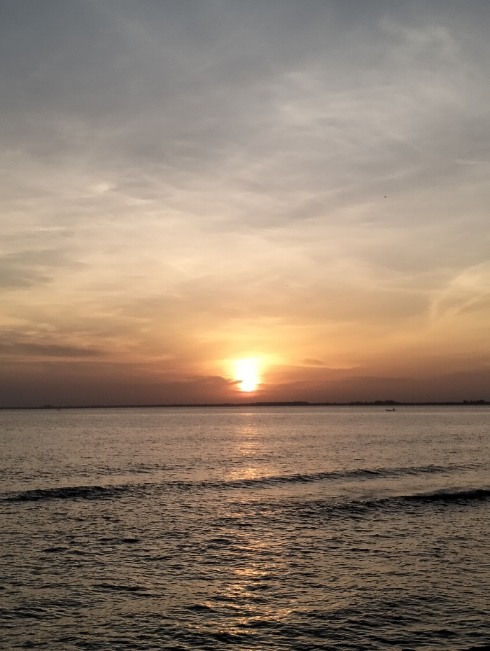 A serene seascape captures a sunset. The sun, partially obscured by a thin cloud layer, casts a warm, golden glow across the water. Subtle waves ripple towards the foreground, reflecting the light. A faint horizon line separates the sea from the sky, transitioning from a soft orange hue near the sun to muted grays above. A tiny boat is silhouetted on the horizon.