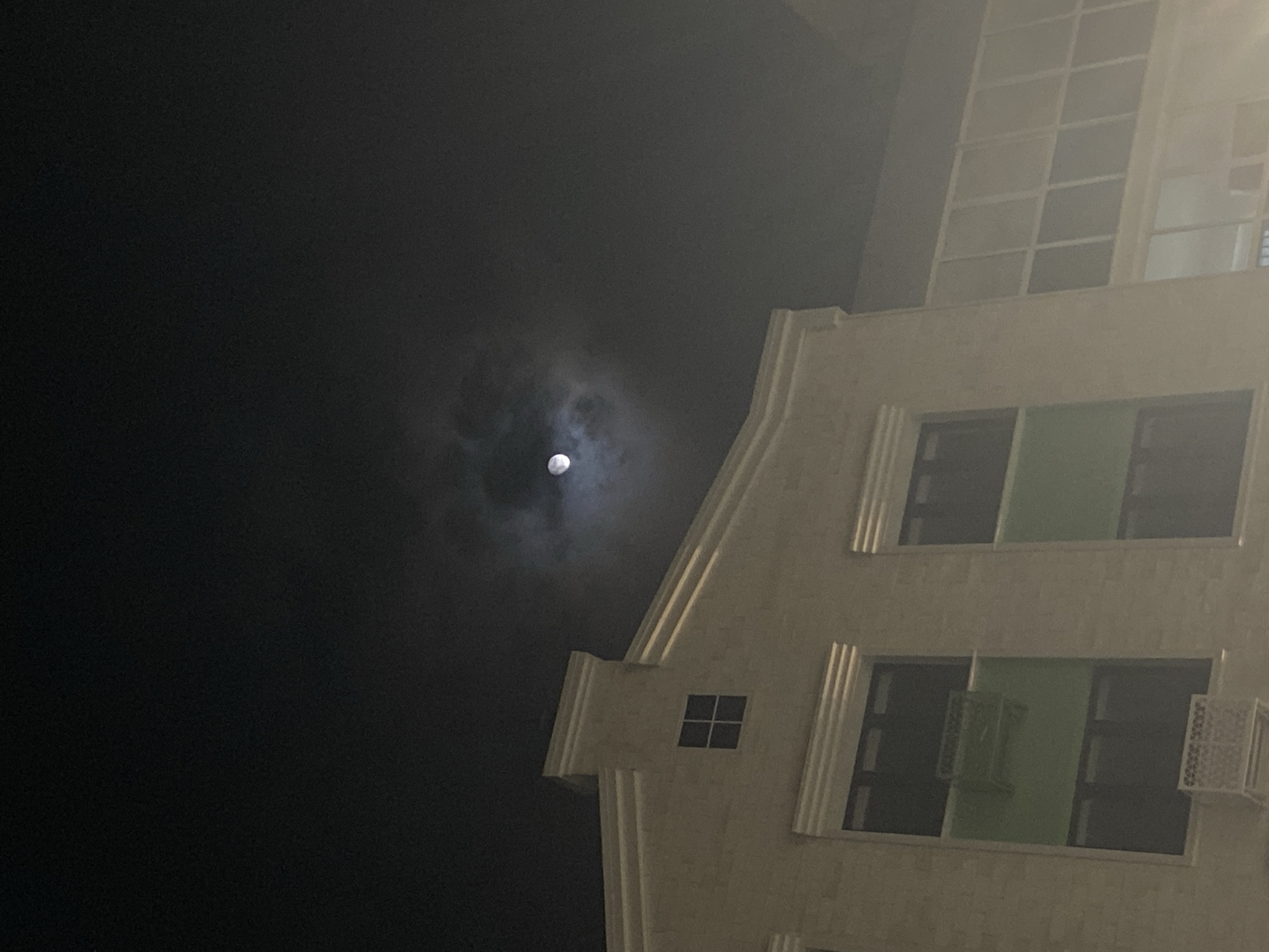 The image depicts a low-angle night shot of a building with the moon visible in the sky. The building is light-colored and appears to be multi-story, featuring rectangular windows and vertical architectural elements. The sky is dark, with the moon partially obscured by clouds, creating a halo effect around it. There are no people visible in the image. The location in the city of Chemoshur, Russia, is not discernible from the image itself.