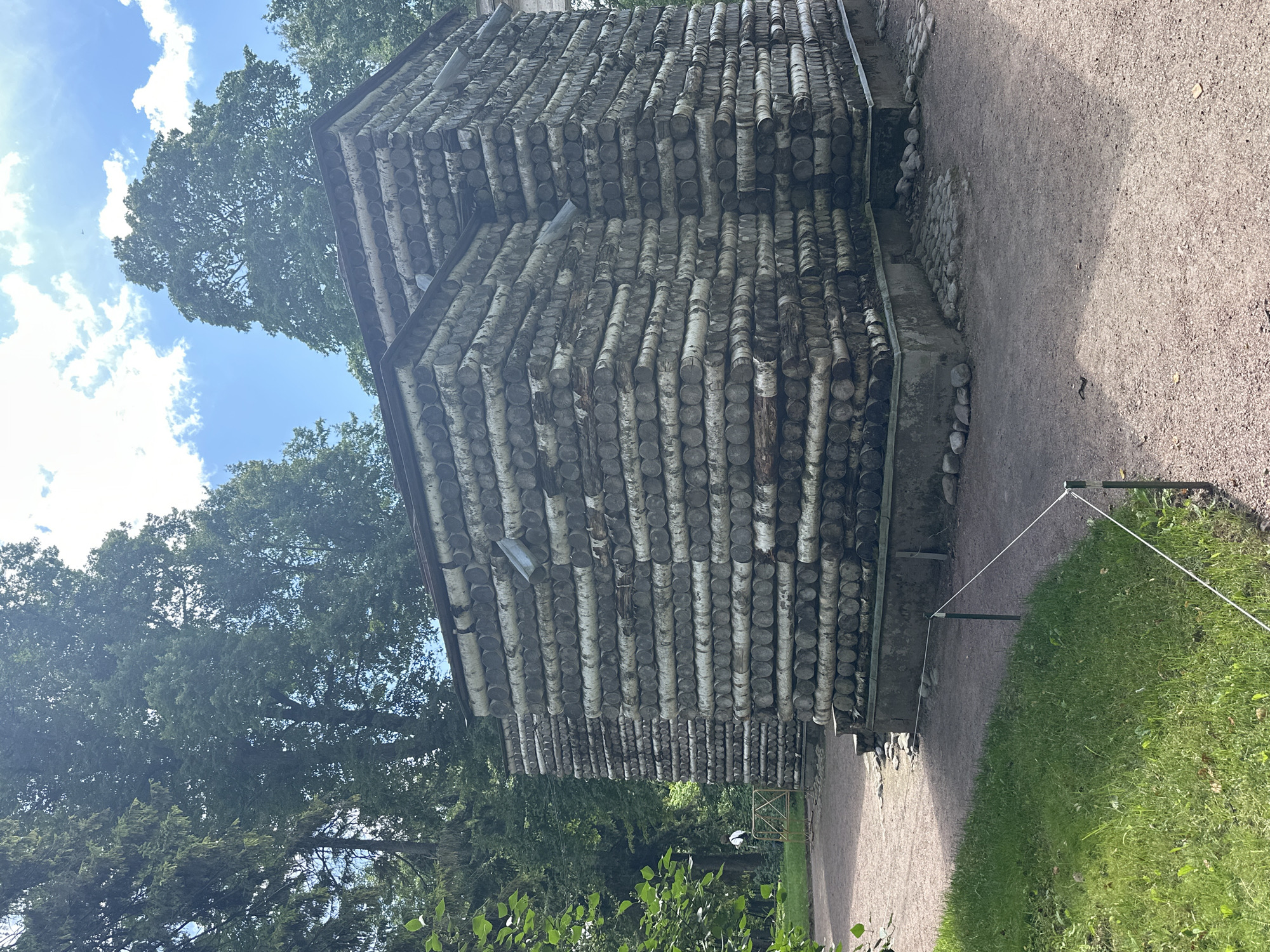 The image shows a building constructed from horizontal logs of various sizes, appearing to be birch logs due to their light color and dark bark striations. The building has a unique, rustic architectural style and is situated in a park-like environment with a gravel path leading up to it and green grass on the left side. Tall trees with dense green foliage surround the structure. A subtle fence made of thin white rope and thin poles delineates an area in front of the building. The sky above is blue with scattered white clouds. This structure is located in the Gatchina Palace and Park Museum-Reserve in Gatchina, Russia, and is known as the Birch House or "Beryozovy Dom". The building appears to be uninhabited in the image.