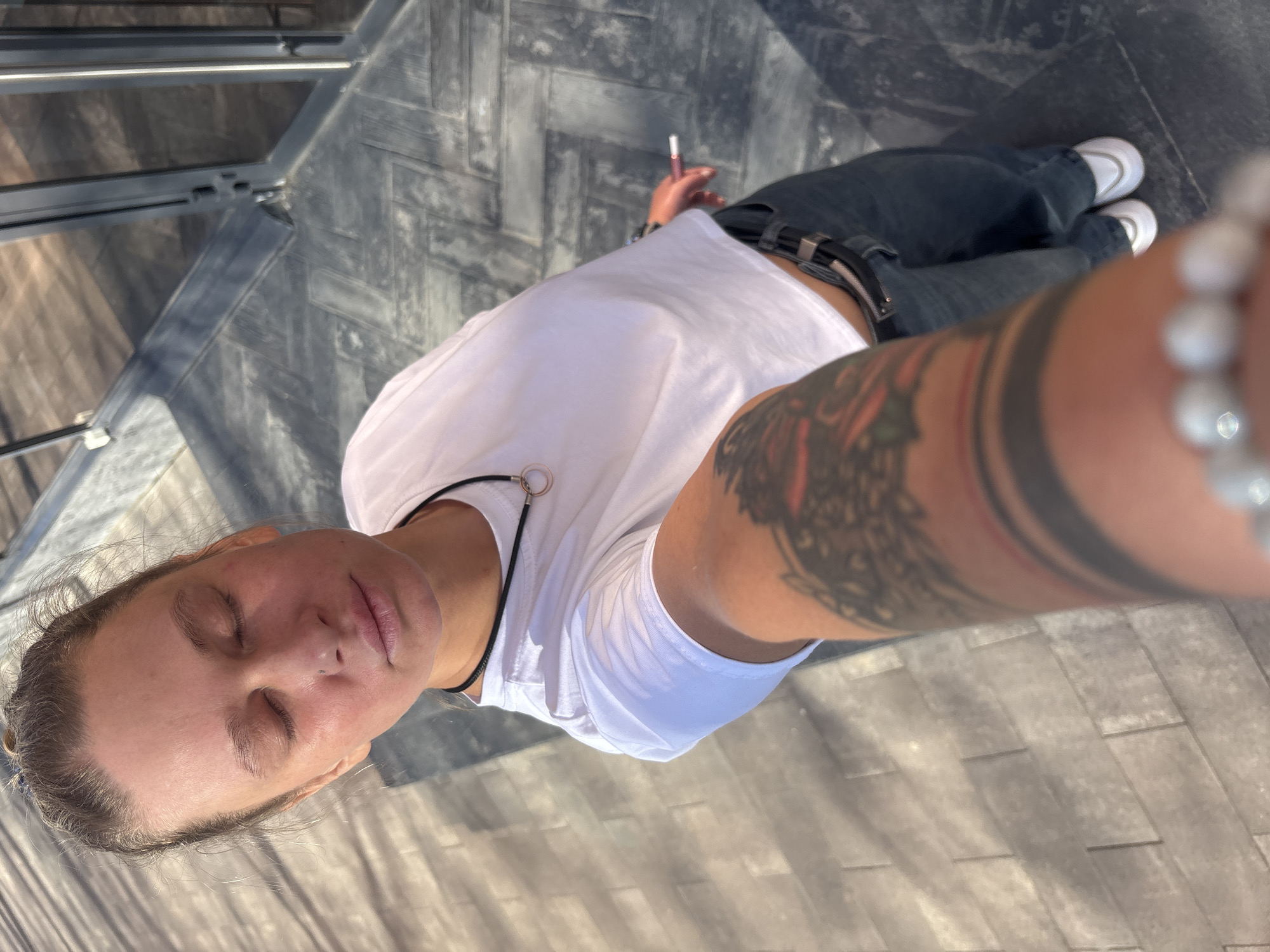 A top-down shot depicts a person with closed eyes, fair skin, and light brown hair. They are wearing a white t-shirt, black pants with a black belt, and white sneakers. The person has tattoos on their right arm and is holding a vape pen in their left hand. The background consists of gray paving stones or tiles arranged in a herringbone pattern, with a metallic structure visible in the upper left corner of the frame. The location is potentially the city of Perm, Russia.