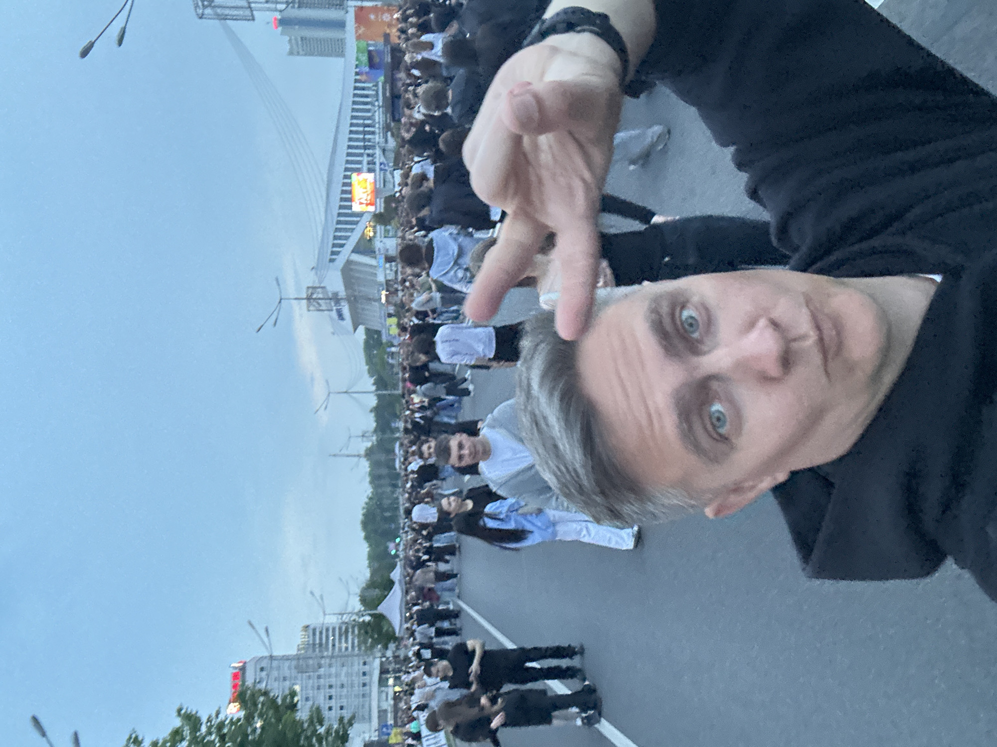 A selfie-style photograph captures a man in the foreground with a large crowd in an urban environment behind him. The man, with grey hair on the sides and blue eyes, wears a black hoodie and makes a two-finger gesture with his left hand towards the camera, partially obscuring the upper right portion of his face. His mouth is slightly pursed.
In the mid-ground and background, a large assembly of people, primarily young adults, occupies a wide asphalt road, indicating a public gathering or event. Many individuals are casually dressed, walking in various directions, some looking towards the camera. White road markings are visible on the asphalt.
The urban environment of Minsk, Belarus, is identifiable by several prominent landmarks. On the left, a tall building with a red illuminated sign that partially reads "АВЕ" (likely part of "AVENUE" or "AVENUE HOTEL", possibly the Hotel Belarus) stands against the skyline. In the center-right background, the distinct white and blue architectural structure of the **Minsk Sports Palace (Дворец спорта)** is clearly visible, featuring a large illuminated screen displaying colorful content. Behind and to the right of the Sports Palace, the cables of the **Pobediteley Avenue bridge** stretch across the sky. Another tall building with a reddish glow at its apex is visible further to the right. The sky is overcast and bright, suggesting late afternoon or early evening. Streetlights are visible throughout the background.