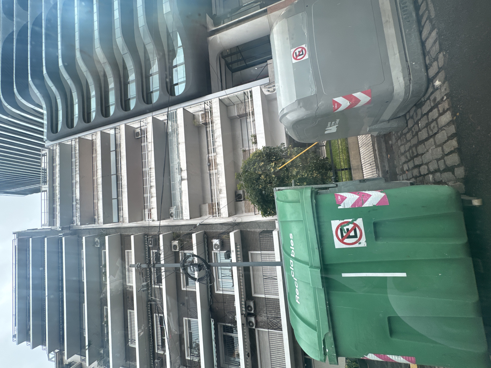 The image depicts a street scene in Buenos Aires, Argentina, with a focus on urban infrastructure and architecture. In the foreground are two large waste containers, one gray and one green. The green container is labeled "Recicla bles", indicating it's for recyclable materials. Both containers have stickers with a symbol inside a red circle with a slash through it. The street surface is paved with cobblestones.
Behind the waste containers, there's a view of several buildings. One building features a modern, curved facade with repeated wave-like elements. Another building has a more traditional design with multiple balconies and rows of windows, and several air conditioning units. Power lines and a utility pole are visible against the backdrop of the buildings. A small tree is present between the buildings and the waste containers.