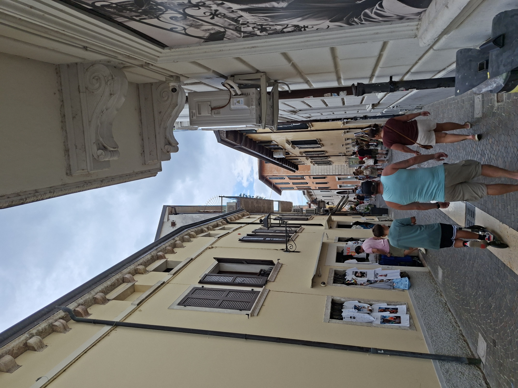 This is a street-level, eye-level, medium shot taken outdoors on a cloudy day in Peschiera del Garda, Italy. The image depicts a narrow, cobblestone street lined with buildings on both sides. The building on the left is a light yellow hue with dark brown shutters on the windows. Several t-shirts are displayed in the lower windows of this building, suggesting it is a shop. The building on the right is white with decorative architectural details, including a large ornate bracket and a mural or large framed picture visible on the upper part of the wall. An air conditioning unit is mounted on the exterior wall of this building.
In the foreground, a young boy is riding a scooter down the street. He is wearing a teal t-shirt and dark shorts. Behind him, a man in a teal tank top and khaki shorts is walking, and a woman in a maroon t-shirt and white shorts is walking beside him. Further down the street, other pedestrians are visible, as are more shops. The street appears to be a commercial area, possibly a pedestrian zone. The overall impression is of a lively street scene in a European town.