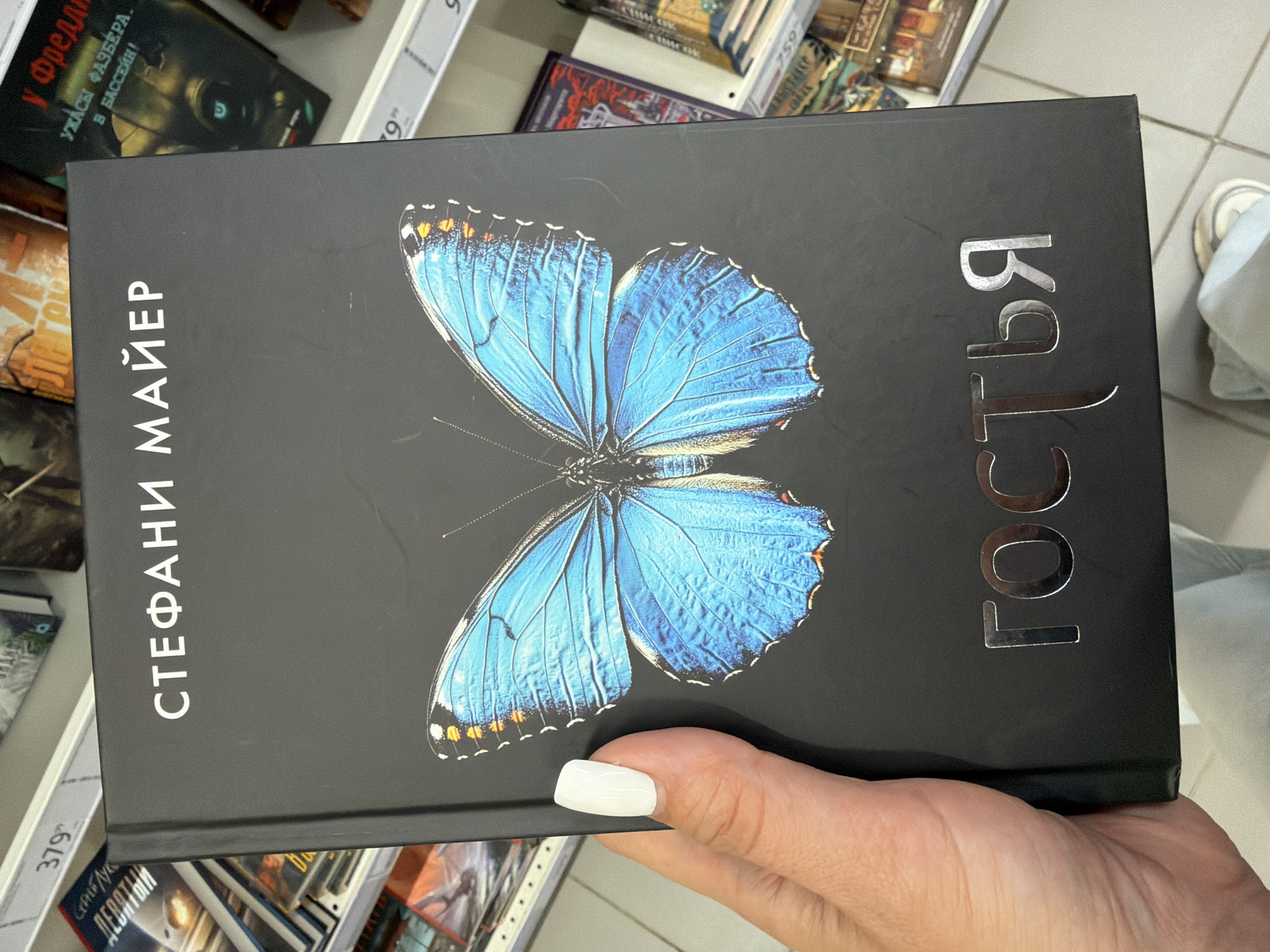 A person holds a book titled "Гостья" by Стефани Майер, featuring a blue butterfly on the cover. The book appears to be in a bookstore, given the presence of other books on shelves in the background. The price tag shows "379." The image may have been captured in the city of Unknown, Unknown, but specific location details are indiscernible.