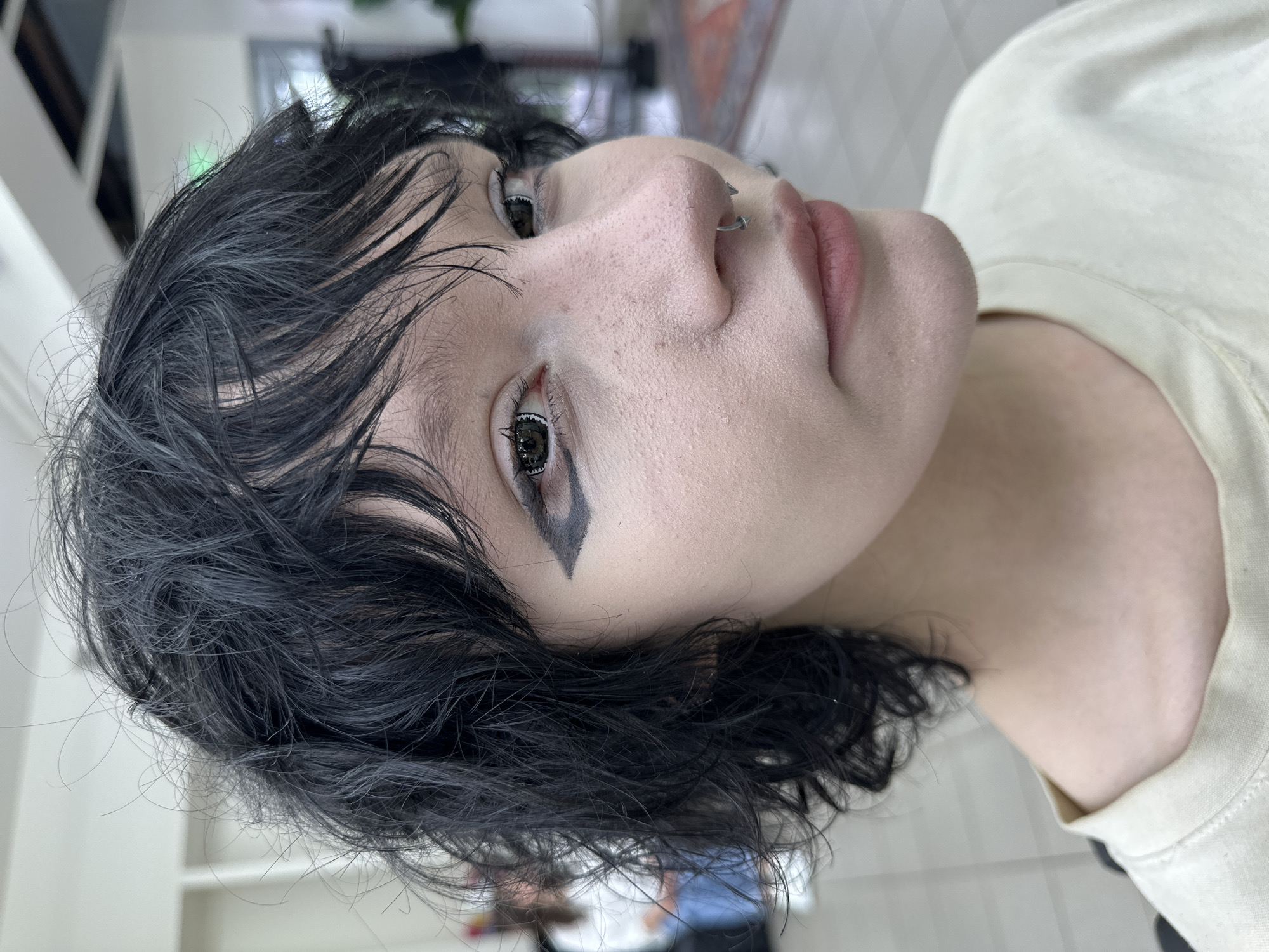 A close-up, angled view of a person's face and upper body, likely taken indoors. The person has fair skin, black hair styled in a shaggy, layered cut, and distinct eye makeup. A silver ring is visible in their left nostril. The person is wearing a light-colored shirt. The background appears to be an interior space with a tiled floor, white walls, and blurred objects. There is no information in the image to indicate that the location is in the city of Chaikovsky, Russia.