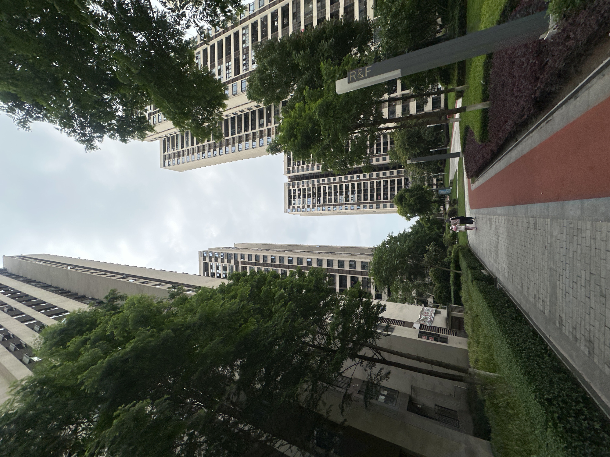 The image shows a street scene in Chongqing High-tech Zone, China. The perspective is looking up towards tall, modern apartment buildings framed by trees on either side. A pedestrian walkway with a red running track borders the buildings. People can be seen walking along the walkway. A sign with "R&F" is visible, likely indicating the name of the real estate development. The sky is overcast. The buildings are high-rise structures with repeating window patterns. The surrounding area includes green landscaping.